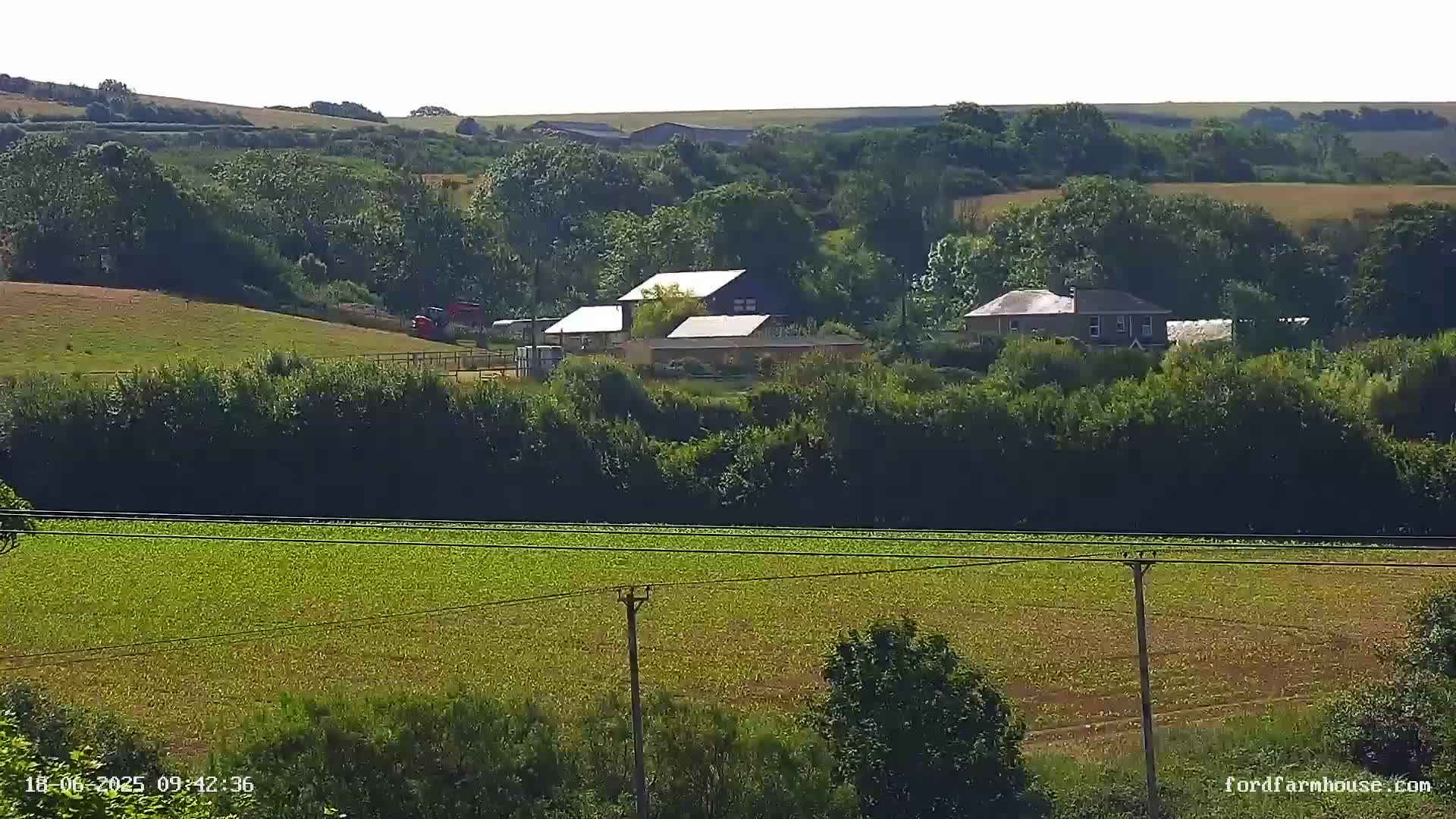A sunny view of a rural landscape shows several buildings nestled amongst green trees and fields, with power lines running across the foreground.