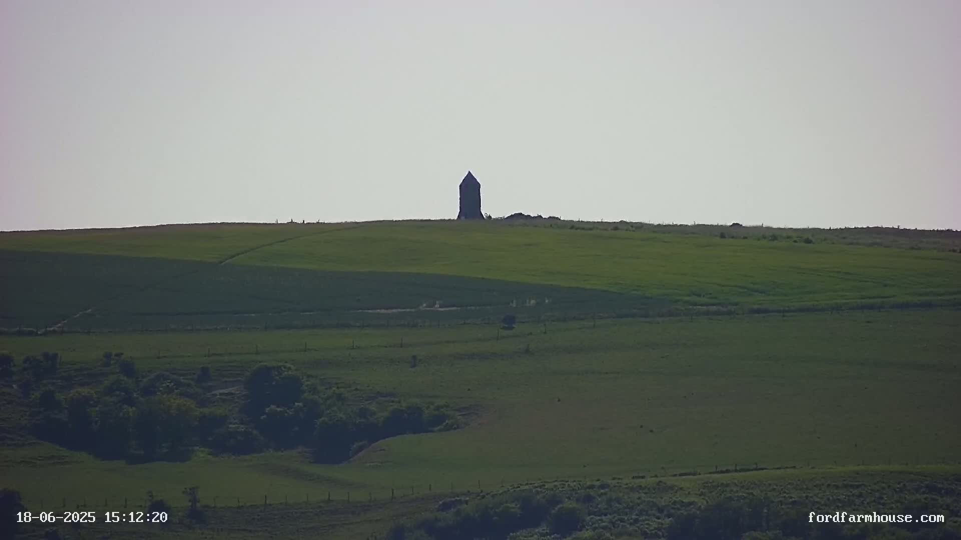 A tall, dark structure stands atop a gently sloping green hill under a light gray sky.