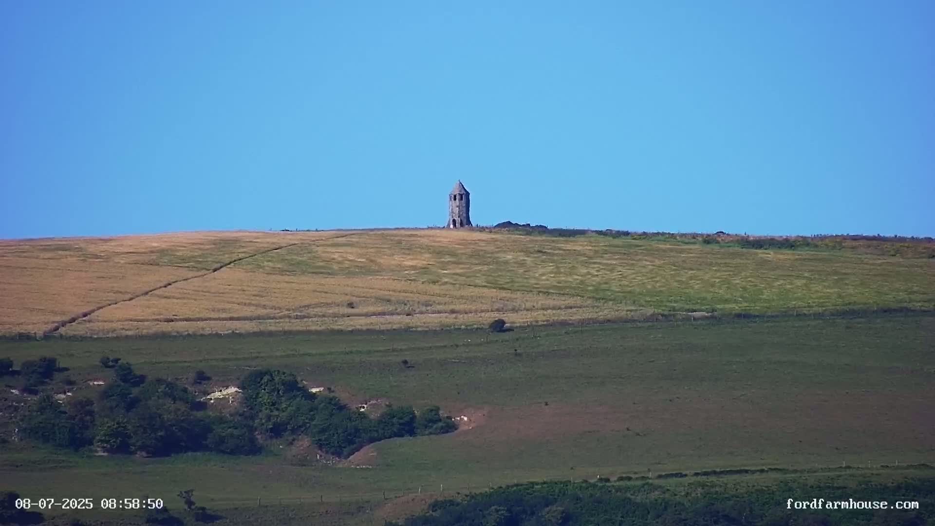 A stone tower stands atop a gently rolling hill under a clear blue sky, with fields of varying shades of green and brown stretching below.