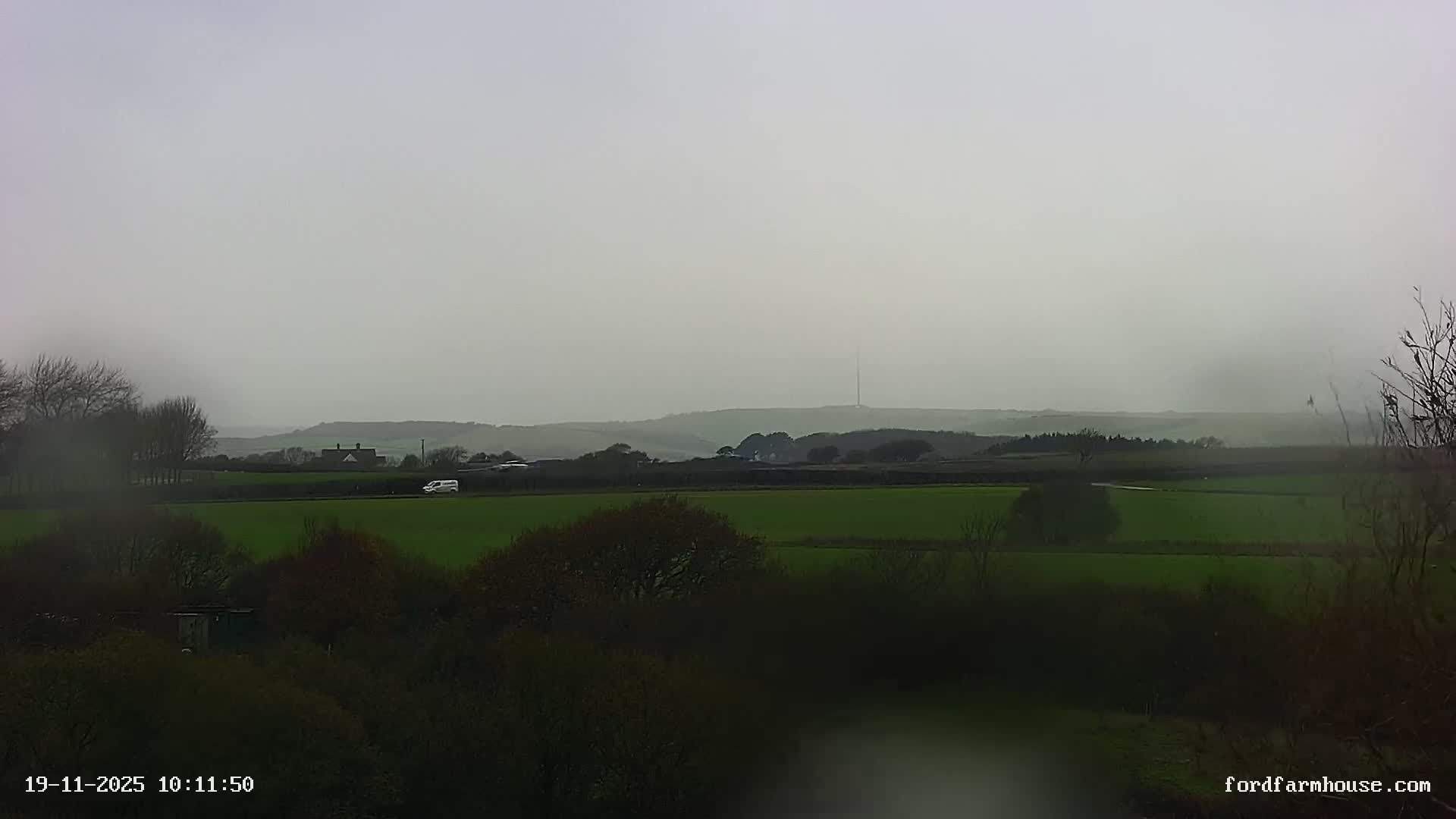 A rural landscape featuring green fields, scattered trees, distant hills, and a white van on a road is viewed under an overcast and rainy sky.