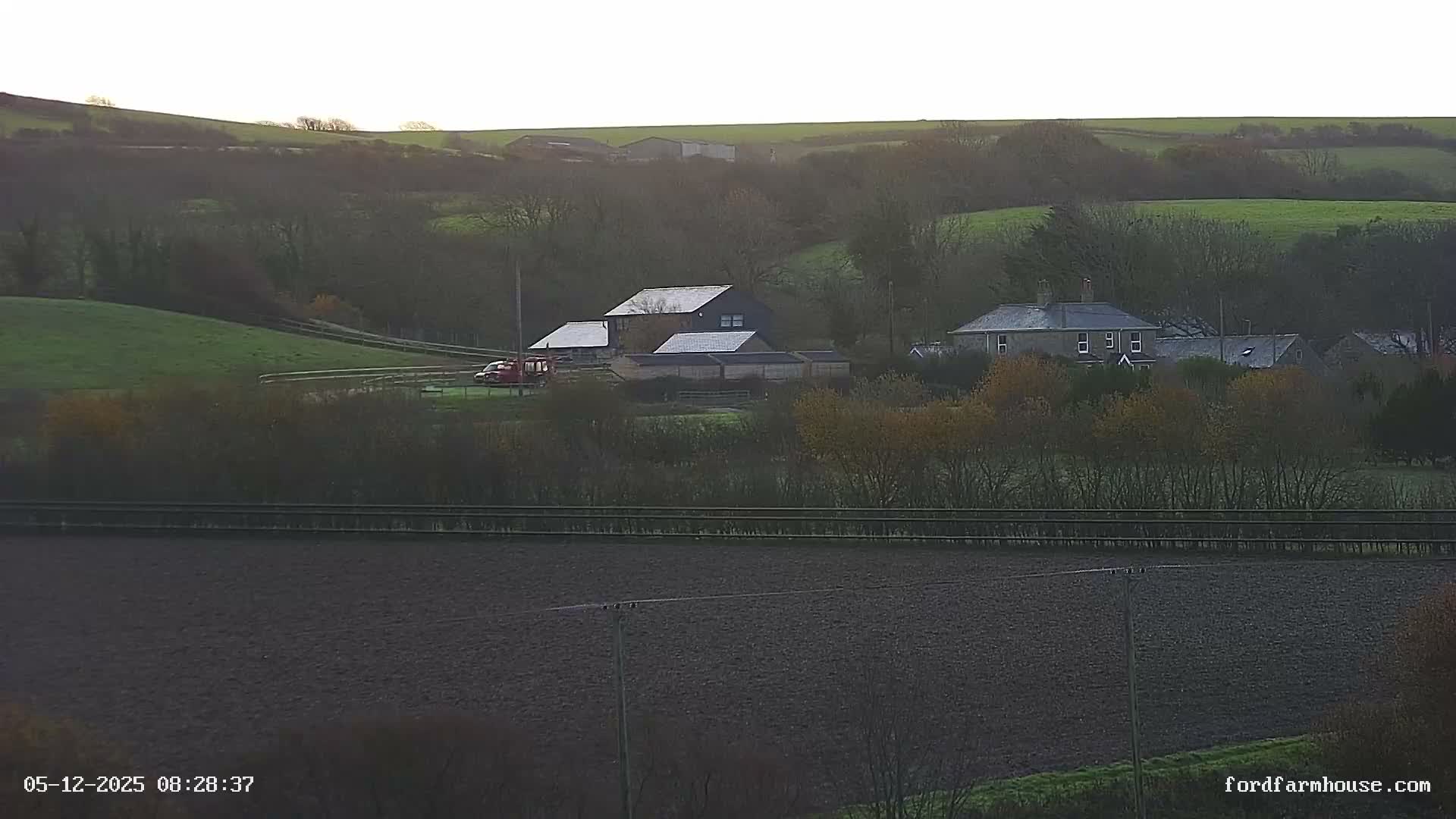 An overcast and cool morning reveals a rural landscape with several houses and outbuildings nestled among rolling green pastures and tilled fields, bordered by trees displaying late autumn foliage, and a red vehicle visible near a fenced track.