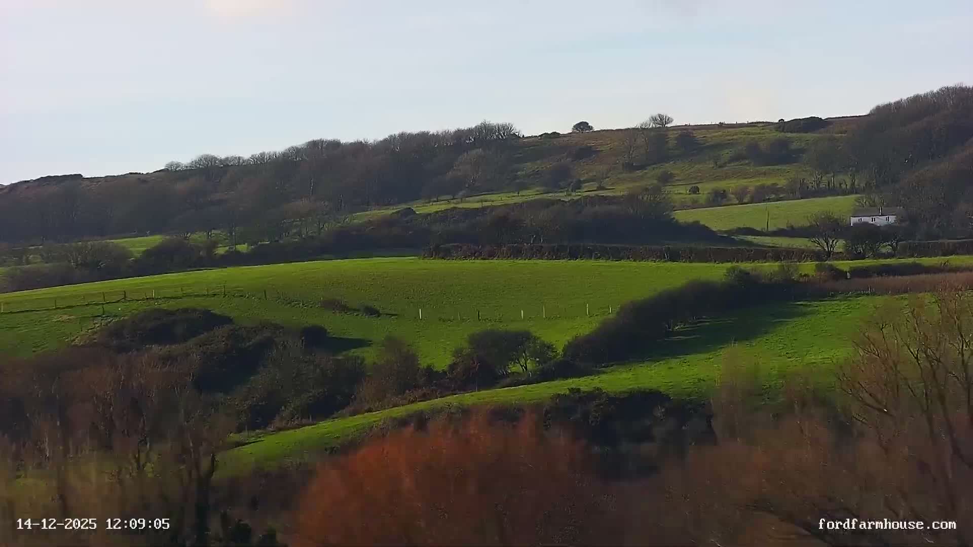 An overcast and cool morning reveals a rural landscape with several houses and outbuildings nestled among rolling green pastures and tilled fields, bordered by trees displaying late autumn foliage, and a red vehicle visible near a fenced track.