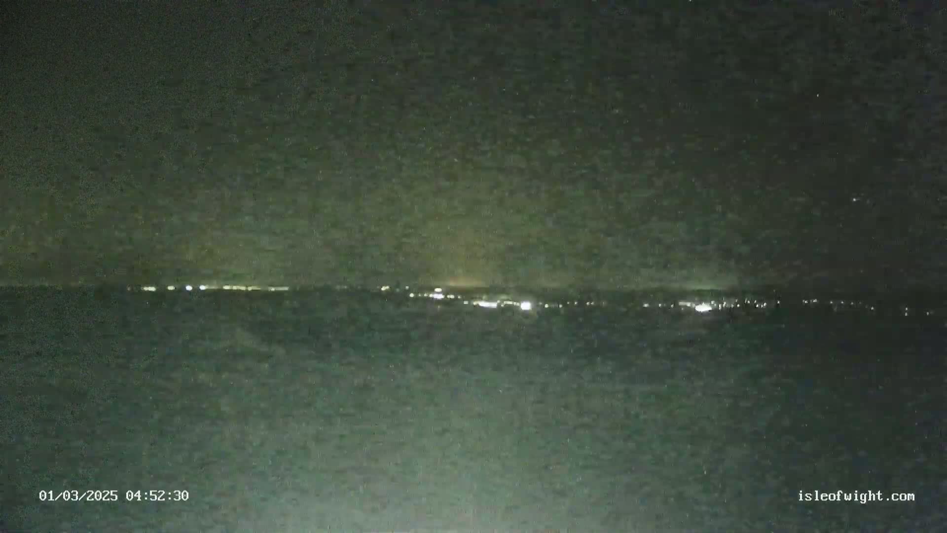 A nighttime long shot shows a dark body of water with a distant line of lights on the horizon under a dark, cloudy sky.