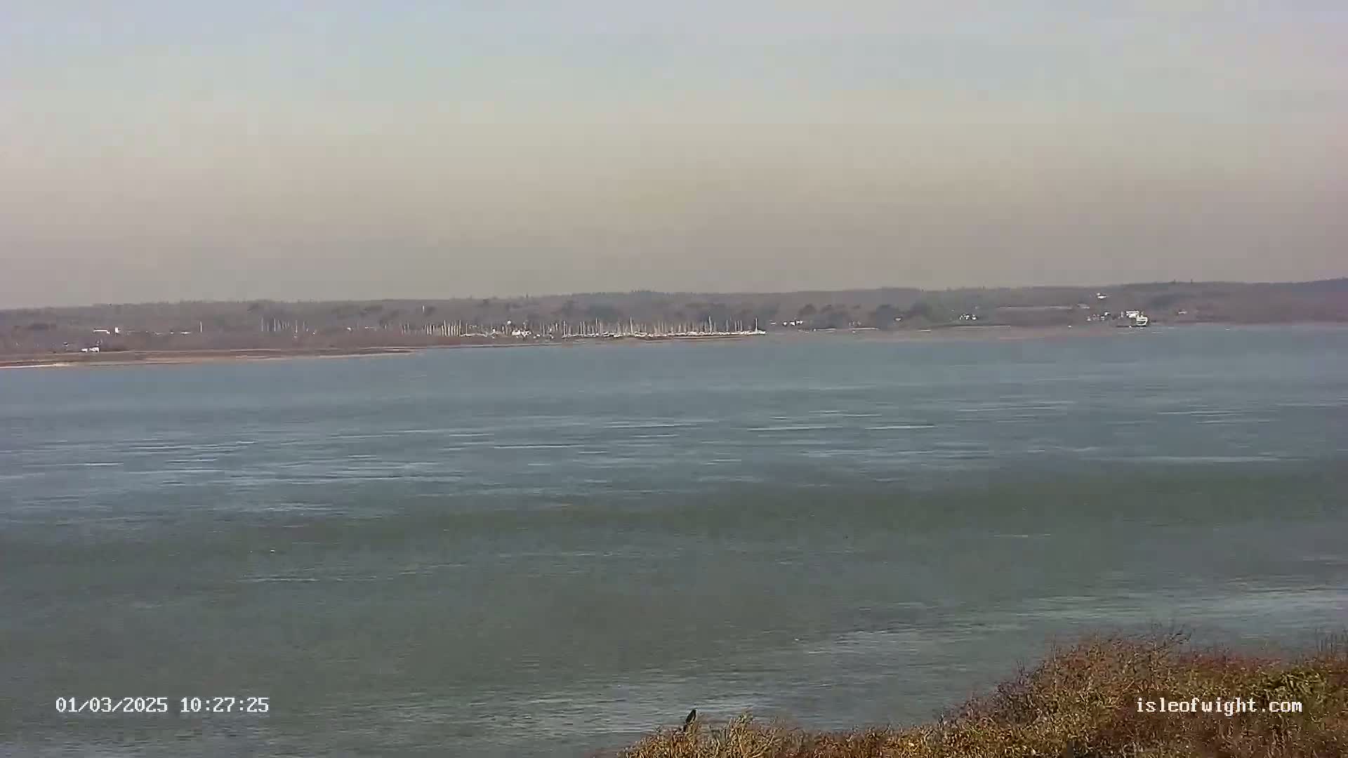A wide, calm body of water stretches to a distant shoreline featuring a collection of boats and a hazy sky.