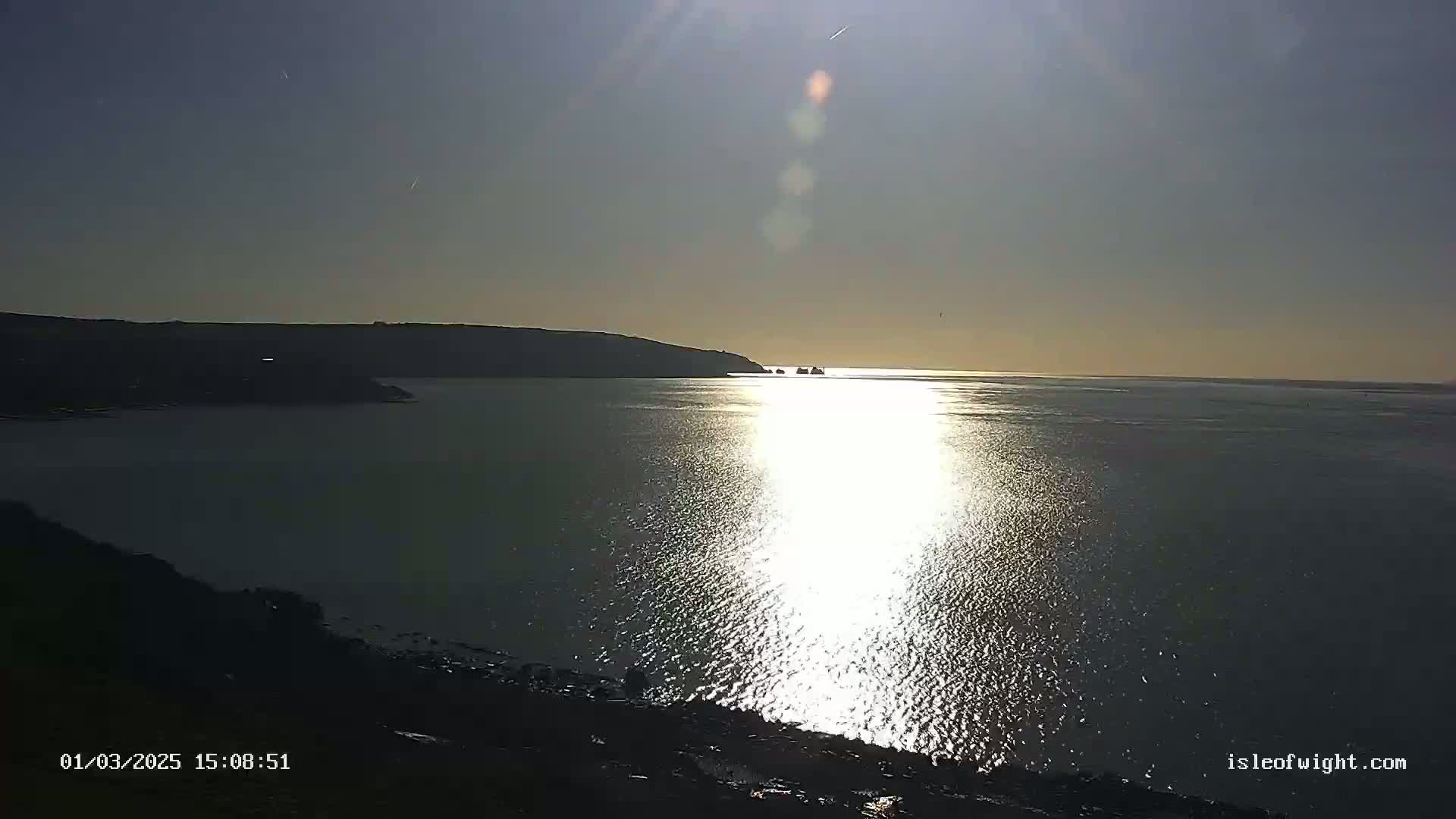 A bright, sunny day shows a calm body of water reflecting sunlight, with a dark landmass in the background and a few small, distant islands visible.