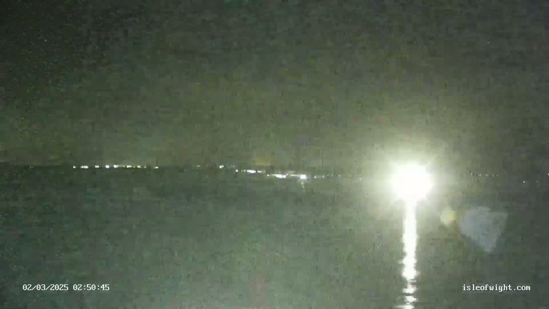 A bright light shines on a dark body of water at night, with a distant line of lights visible on the horizon.