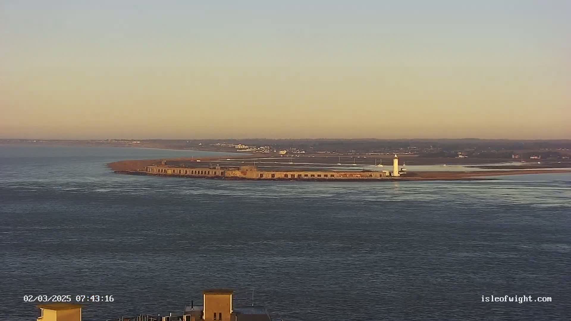 A long, low stone structure with a lighthouse at one end sits in calm water under a pale, clear sky at sunset.