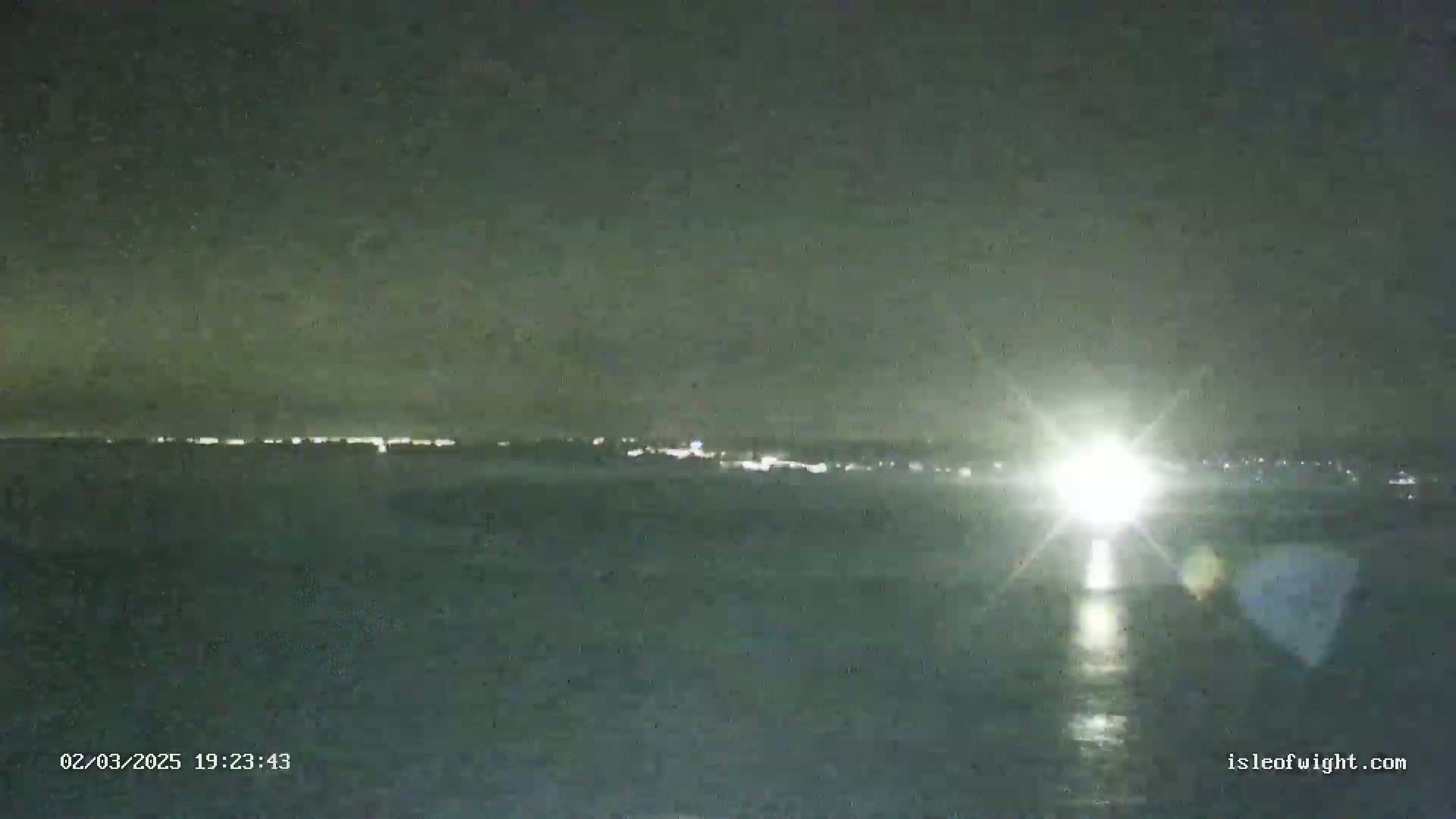 A nighttime view of a body of water with a distant shoreline showing faint lights under a dark, mostly clear sky.