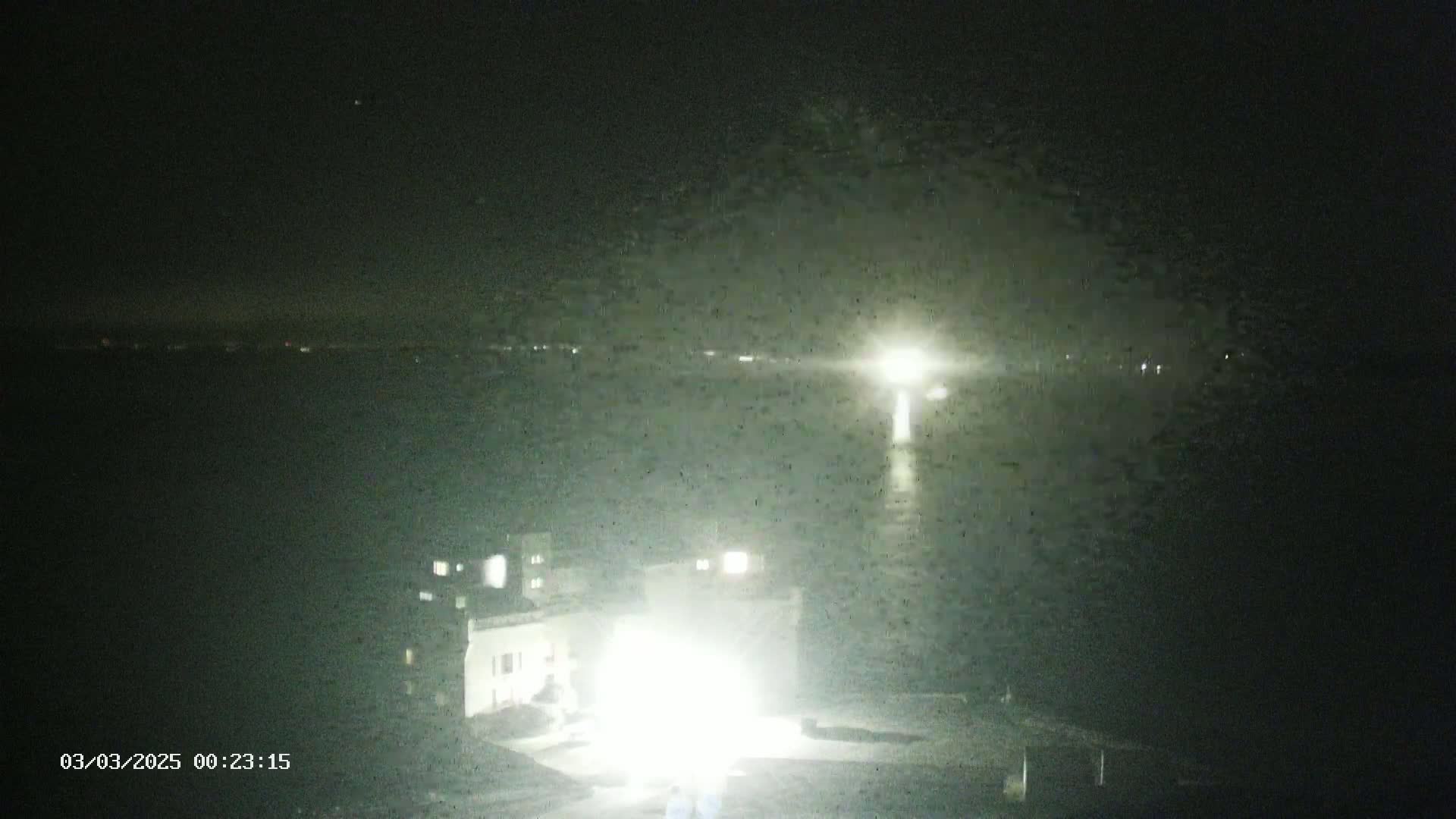 A nighttime, low-light view shows a body of water reflecting light from a distant source, with a brightly lit structure and faintly lit buildings in the foreground.