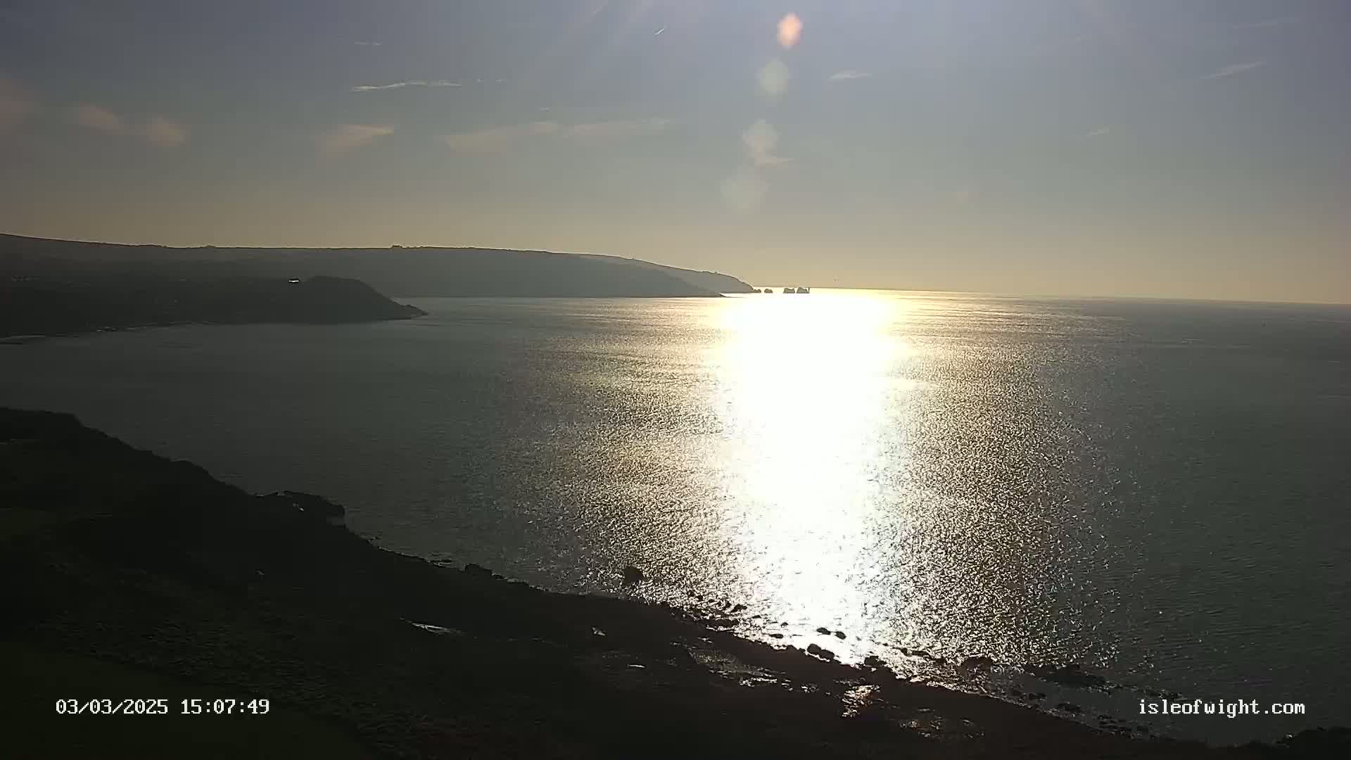 A bright sun shines on a calm sea near a rocky coastline and distant headland under a mostly clear sky.