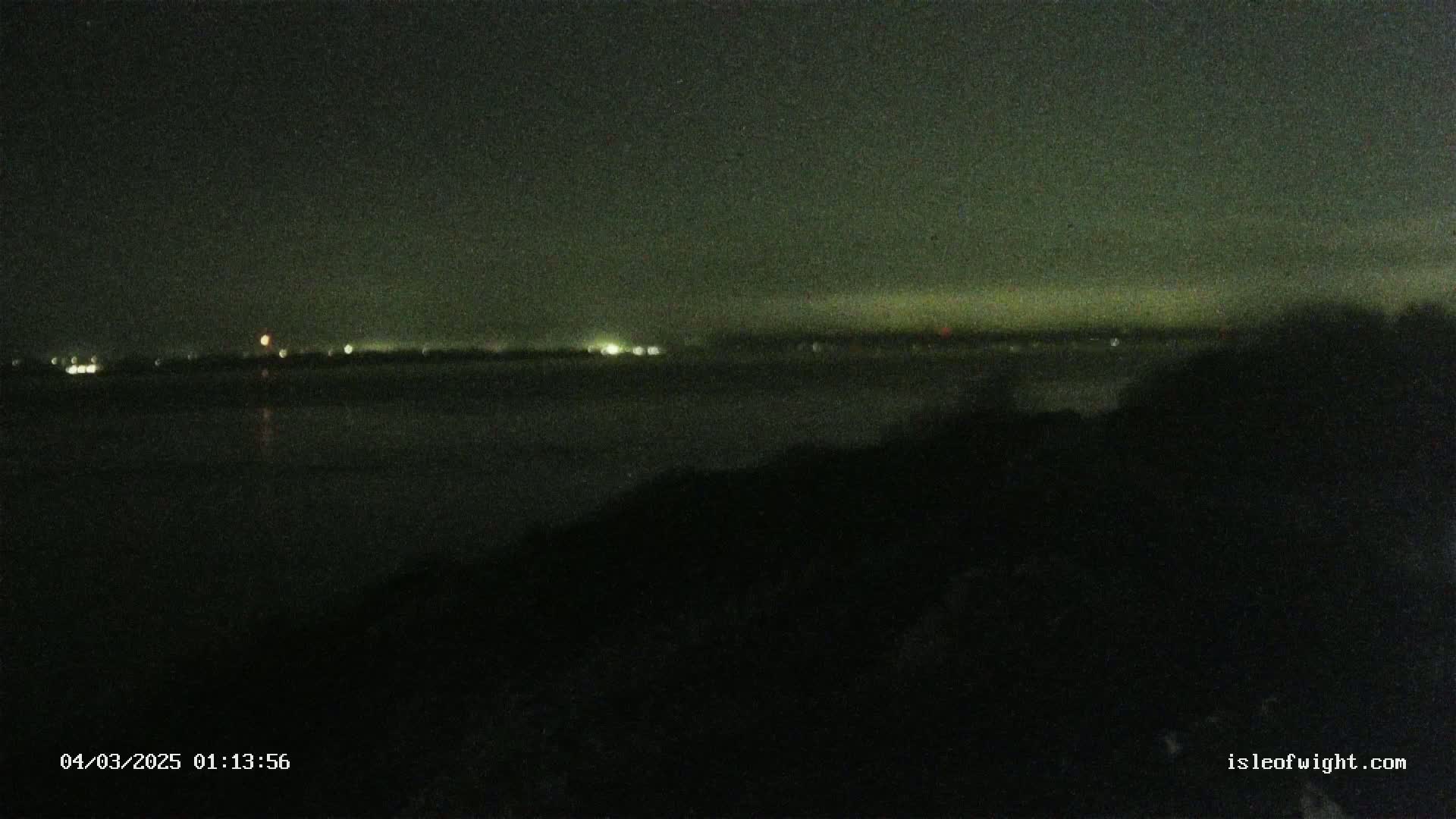 A dark nighttime view of a body of water with faintly visible lights on the opposite shore under a mostly clear sky.