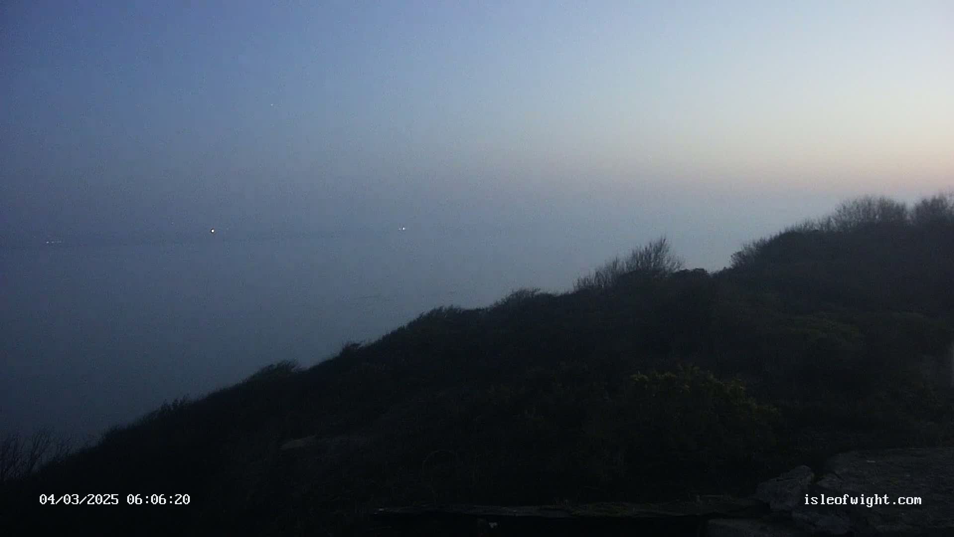 A dark, foggy hillside overlooks a hazy, light-blue body of water under a twilight sky.