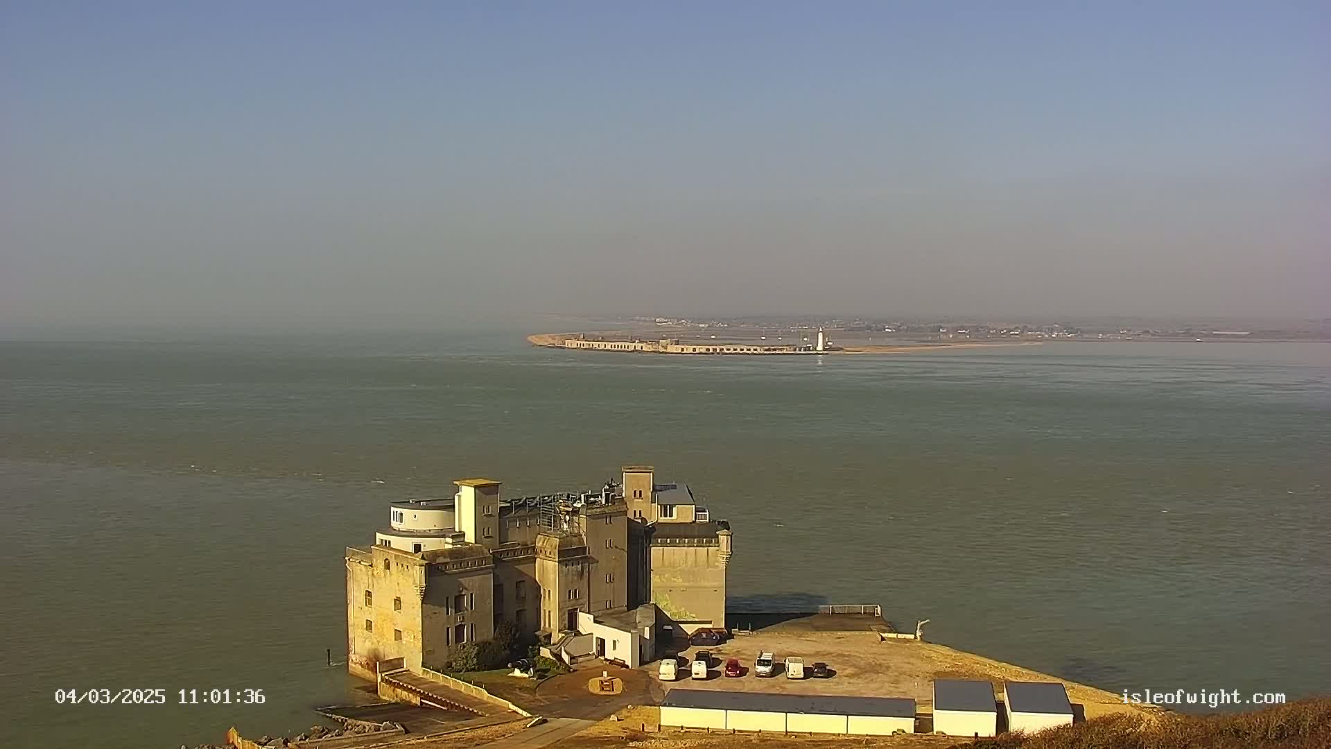 A large, stone building sits on a cliff overlooking a calm body of water, with a distant breakwater and lighthouse visible under a clear, hazy sky.