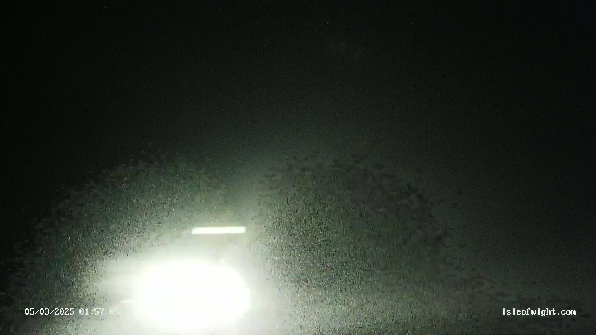 At night, a bright light from a vehicle's headlights illuminates a rain- or snow-covered ground.