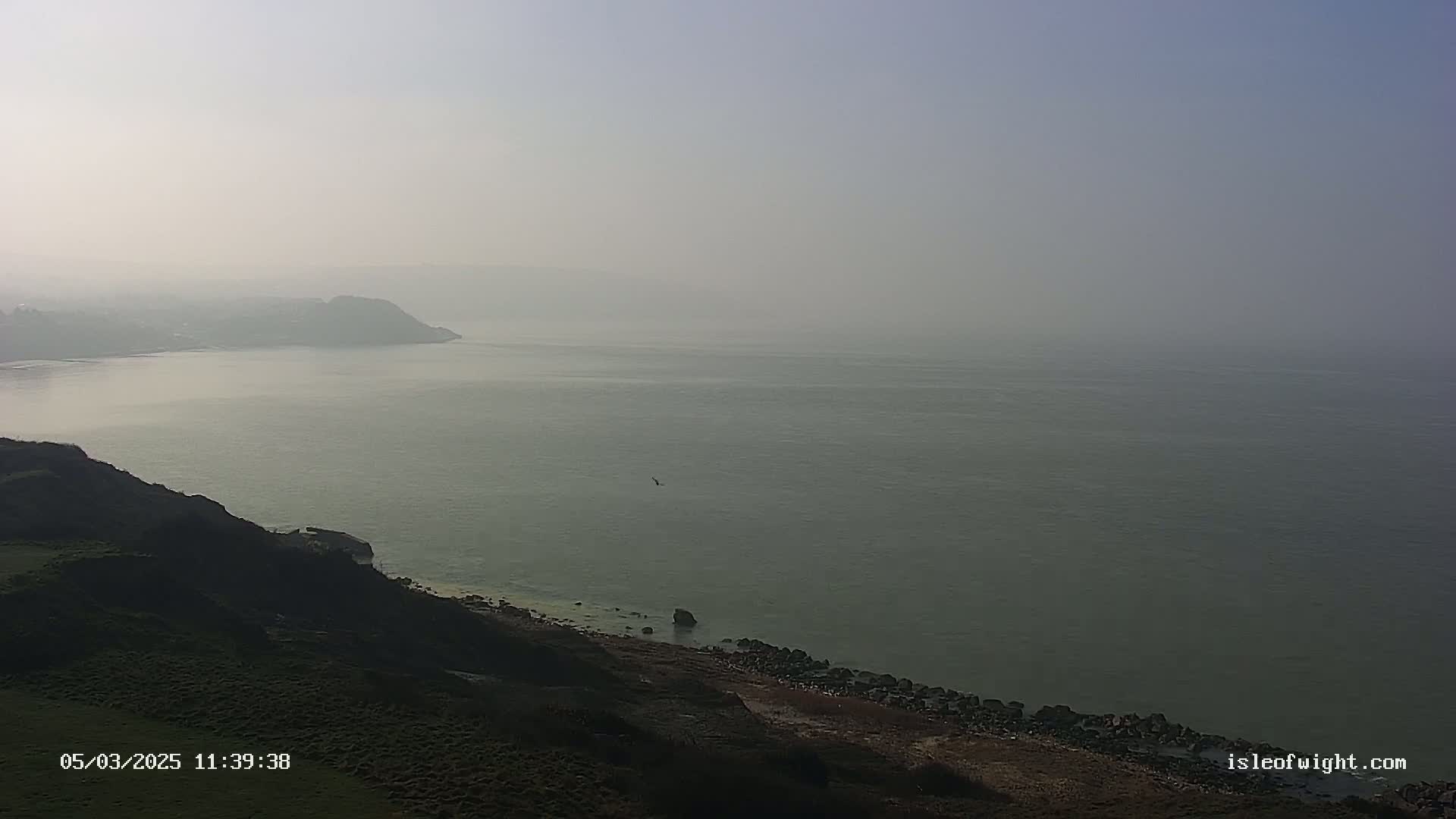 A hazy, overcast day reveals a coastal scene of cliffs, a rocky shoreline, and a calm sea extending to a distant, indistinct landmass.