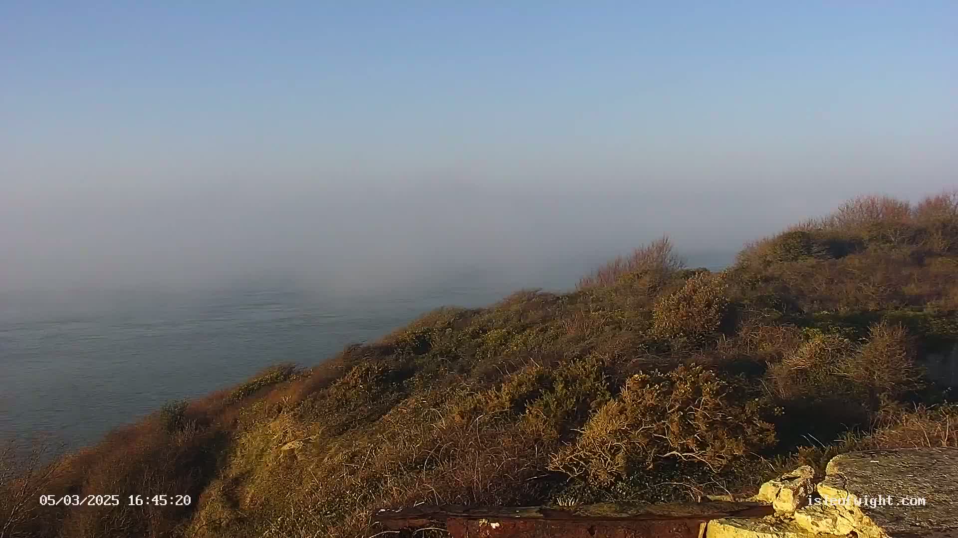 A hazy, calm body of water is visible from a coastal cliff covered in low-lying, brown vegetation.