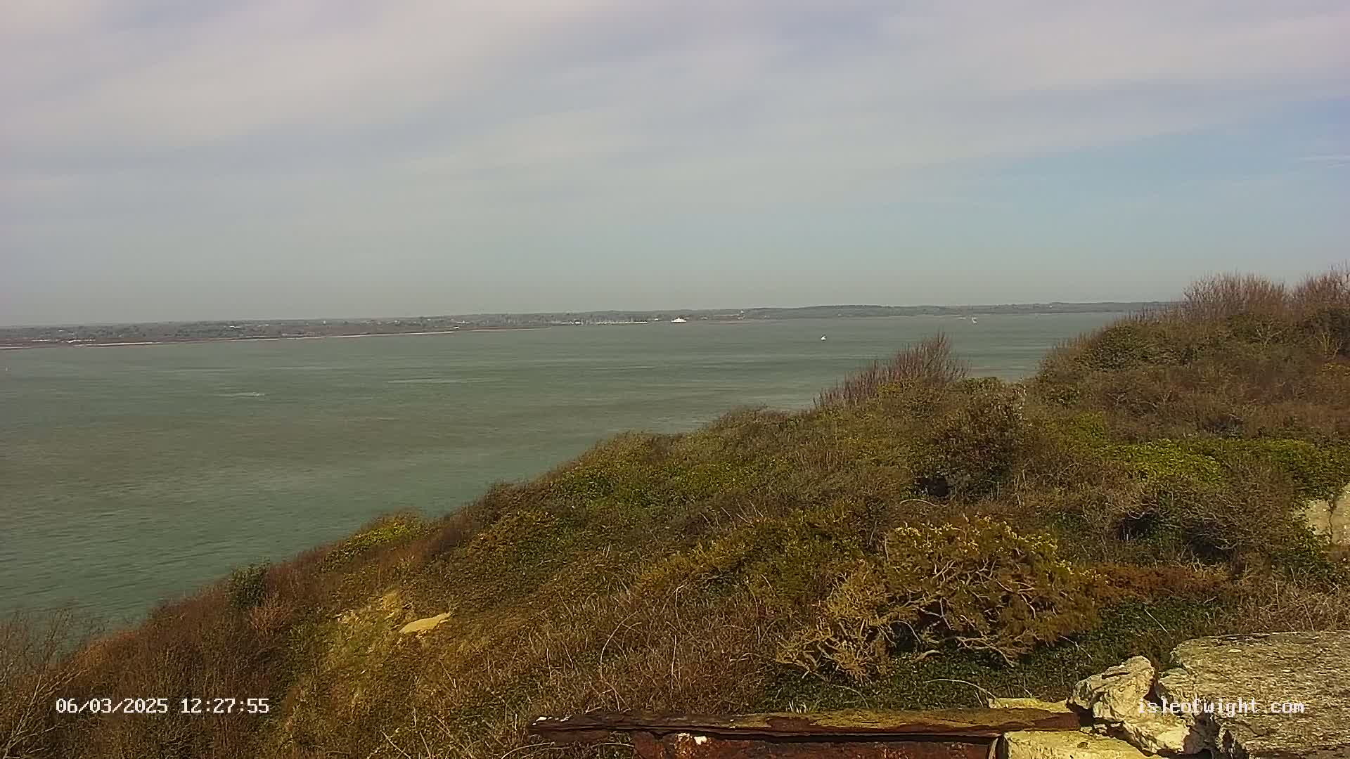 A tranquil coastal scene on a partly cloudy day shows a calm body of water extending to a distant shoreline, viewed from a vantage point atop a hillside covered in low-lying, scrubby vegetation.