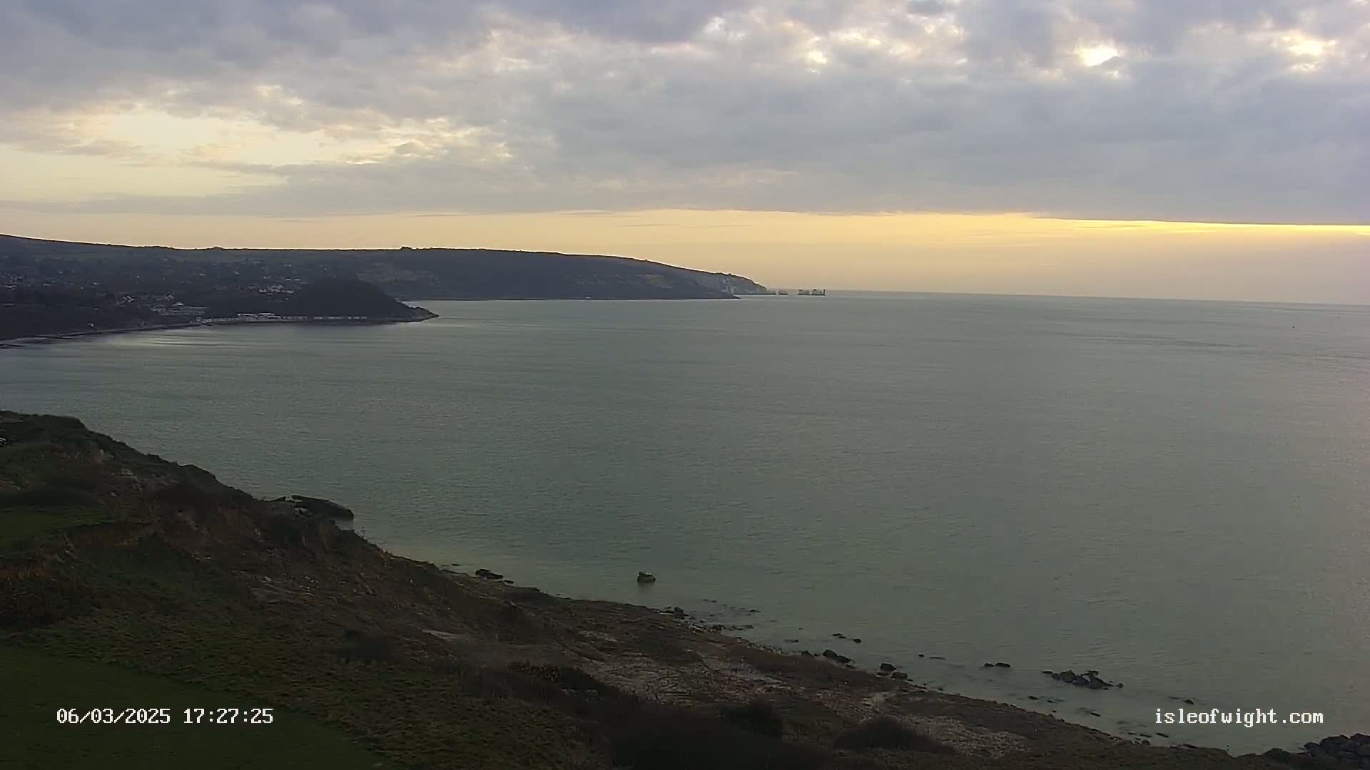 A coastal scene at sunset shows a calm sea, a grassy cliffside, and distant land under a partly cloudy sky.