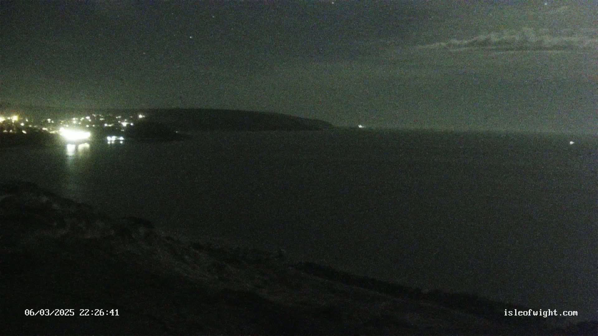 A nighttime view of a dark body of water with a distant shoreline showing faint lights, under a mostly clear sky.