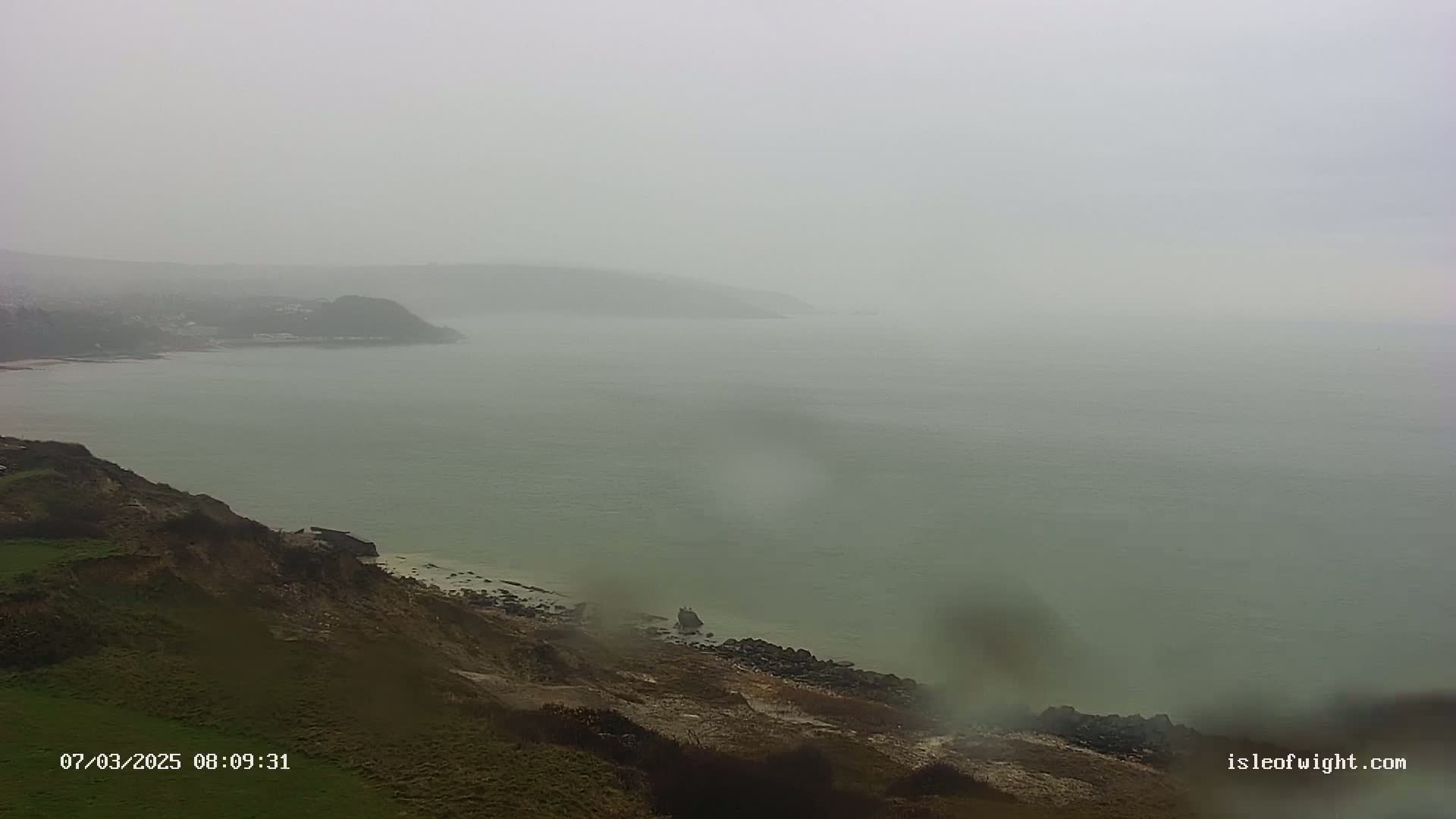 A foggy coastal scene shows a green grassy cliff overlooking a calm, misty sea and distant hills.