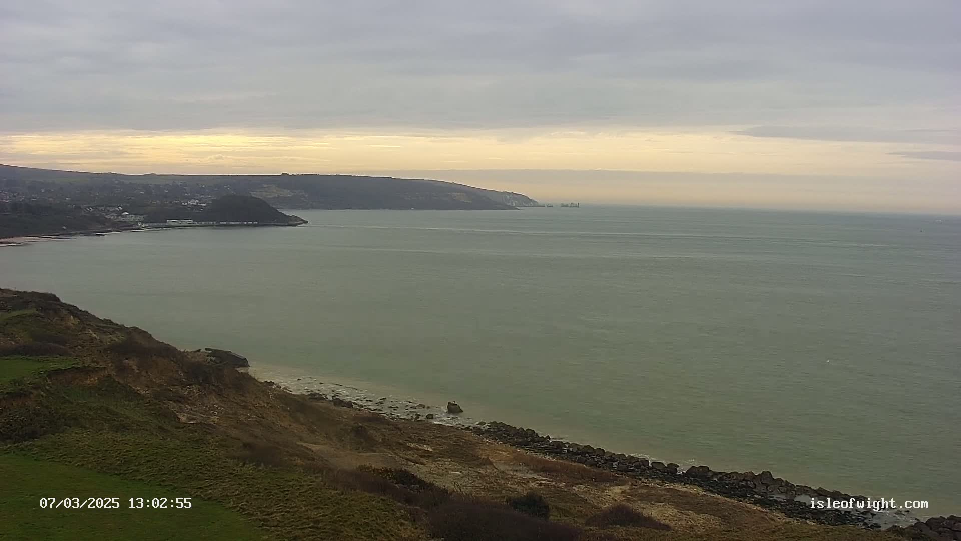 A coastal scene shows a calm, greenish-grey sea under an overcast sky, with a distant town nestled beside cliffs that lead down to a rocky shore.