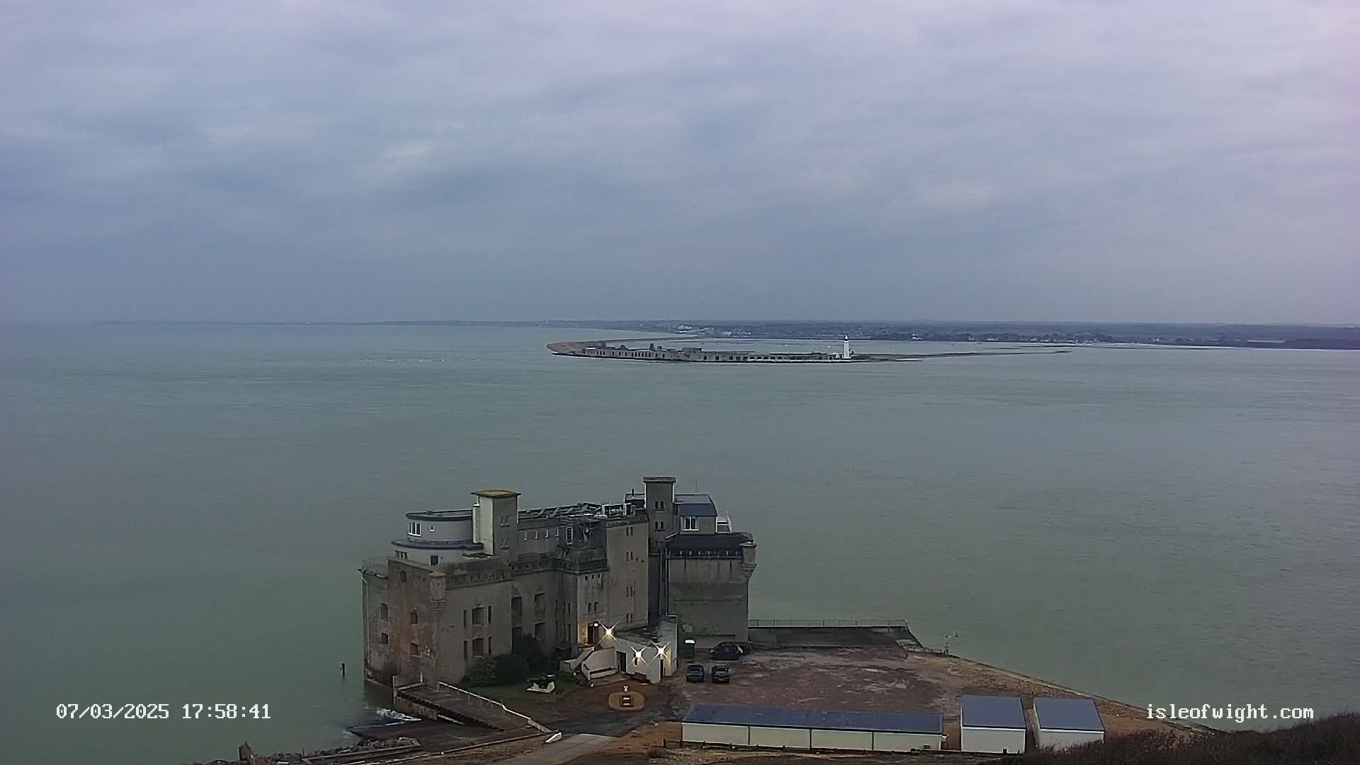 A gray stone castle-like structure sits on a coast overlooking a calm, gray body of water under an overcast sky, with a distant landmass visible in the background.