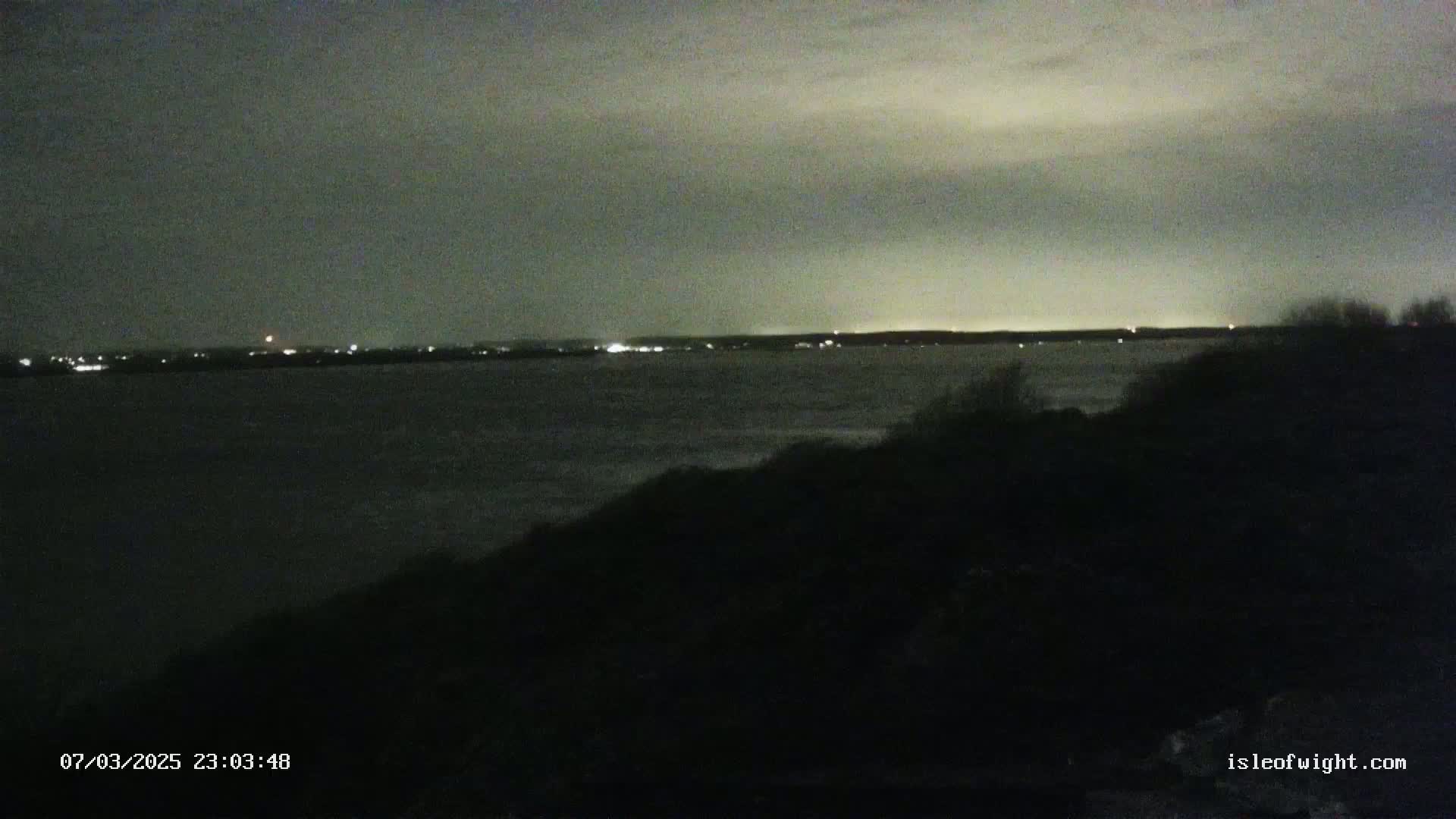 A dark, nighttime view of a body of water with a distant shoreline showing faint lights under a cloudy sky.