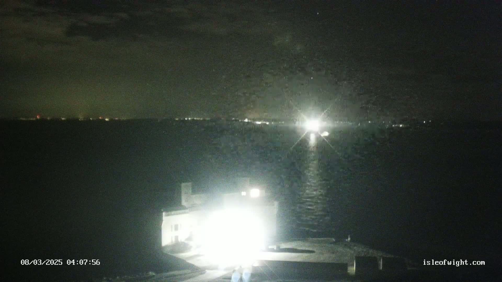A brightly lit building stands near dark water under a mostly clear night sky, with lights visible in the distance across the water.