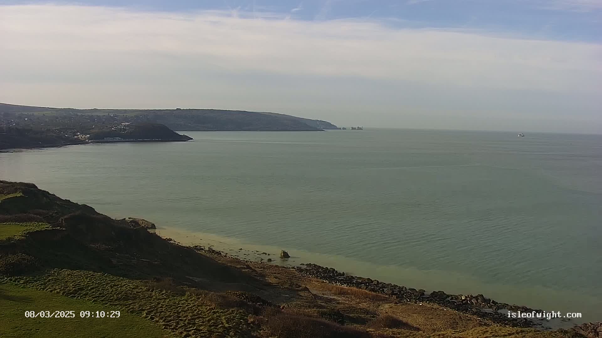 A tranquil coastal scene shows a calm sea meeting a grassy cliffside under a lightly cloudy sky.