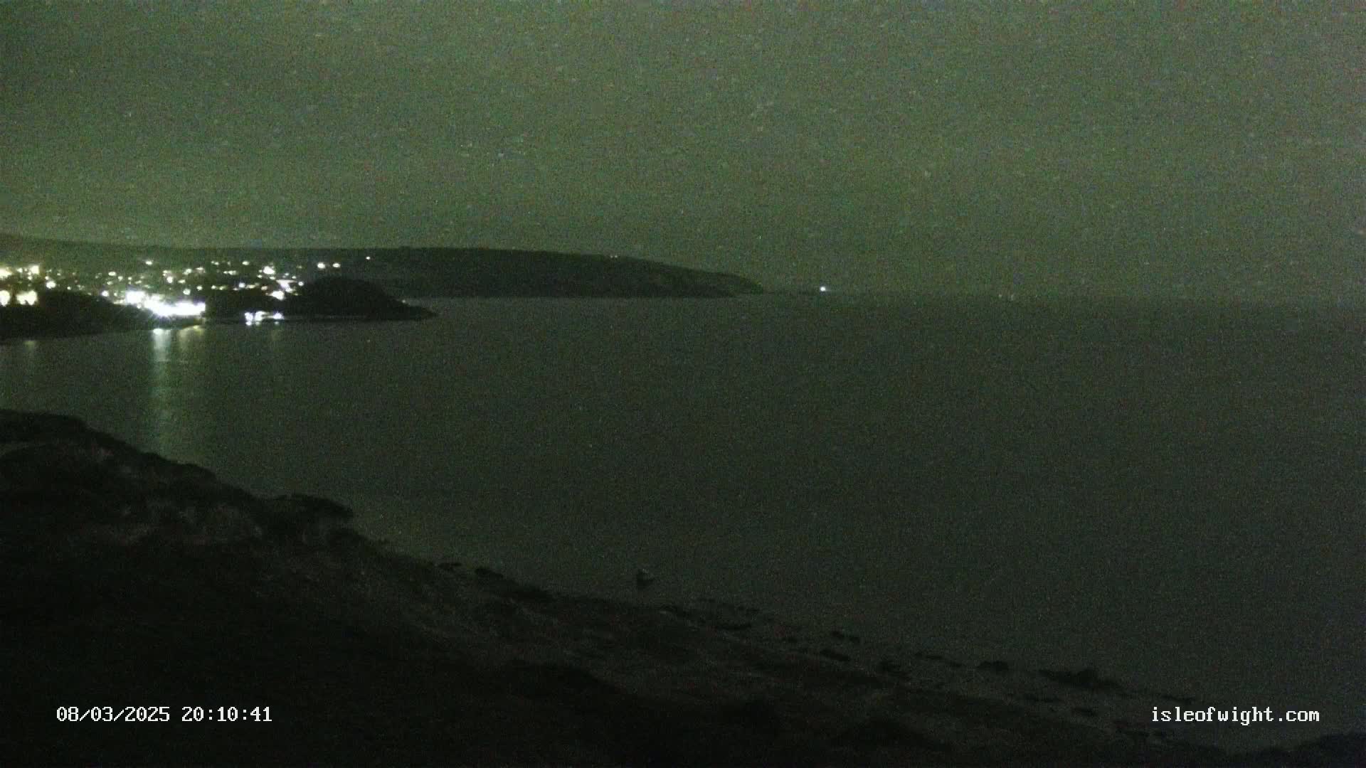 A nighttime view of a dark body of water with a distant shoreline showing faint lights, under a dark, possibly starry sky.