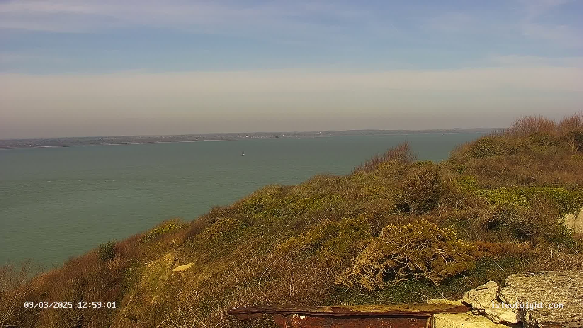 A calm, pale-green body of water is seen from a high vantage point overlooking a hillside covered in low, brown vegetation under a mostly clear, light-blue sky.