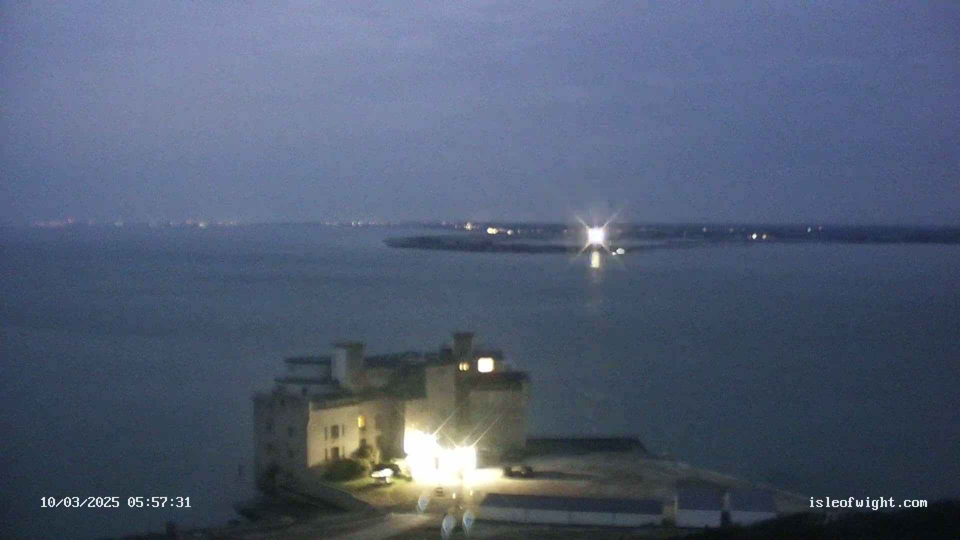A stone building with lights on is situated near a body of water under a dark, twilight sky.