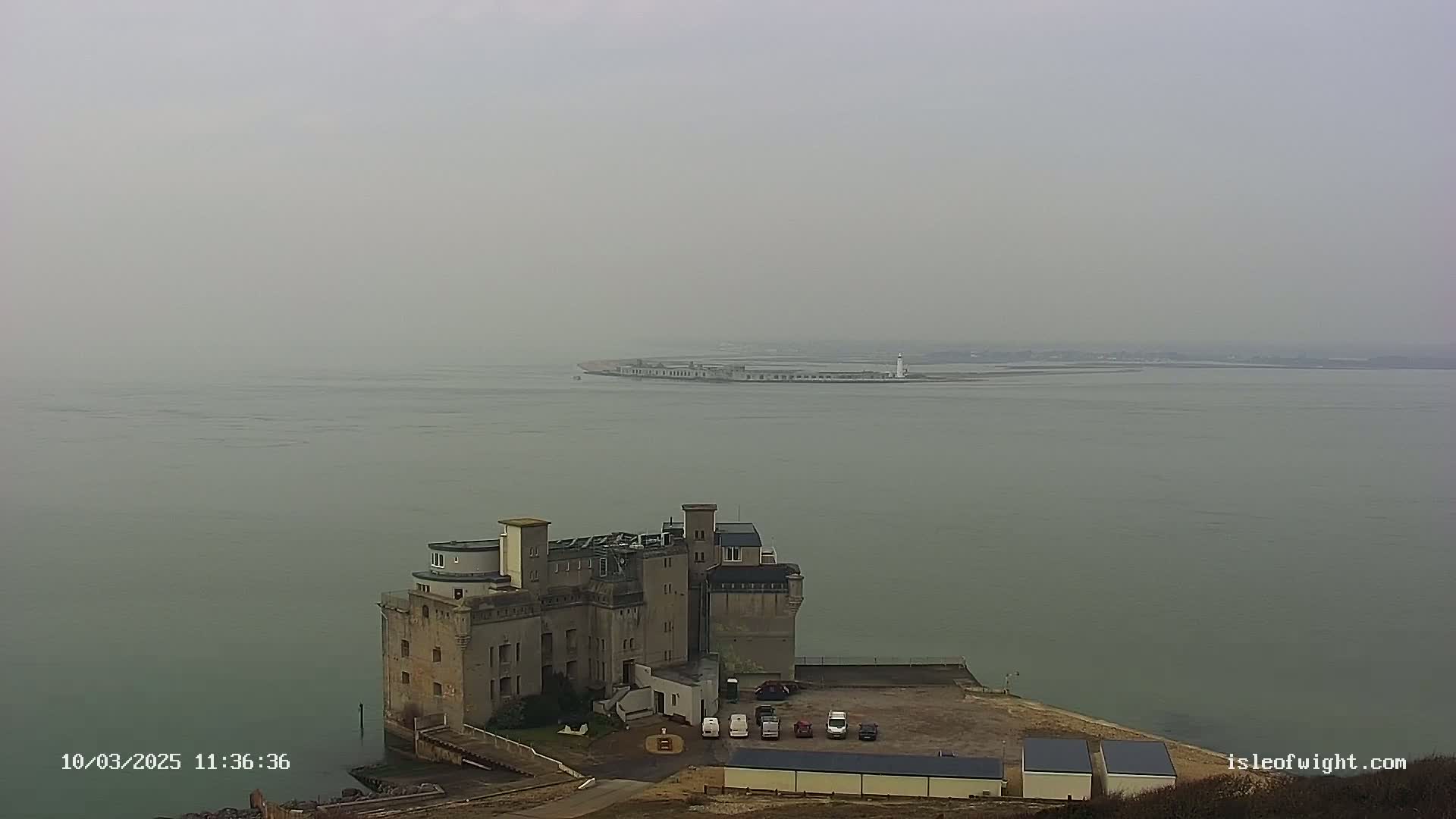 A stone structure resembling a castle sits on the coast near a body of water on a foggy day.