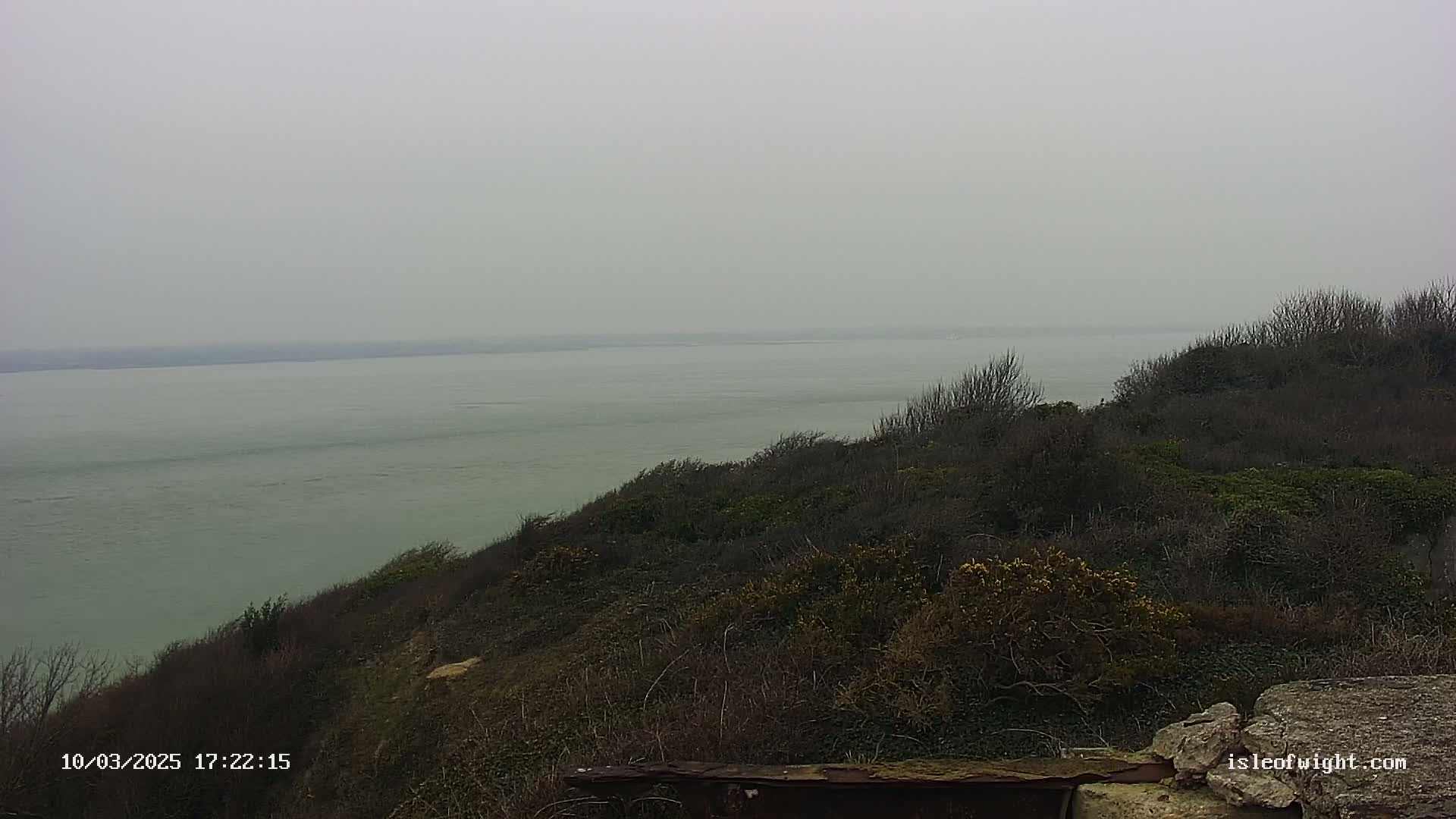 A calm, gray body of water is visible from a hill covered in dark, winter vegetation.