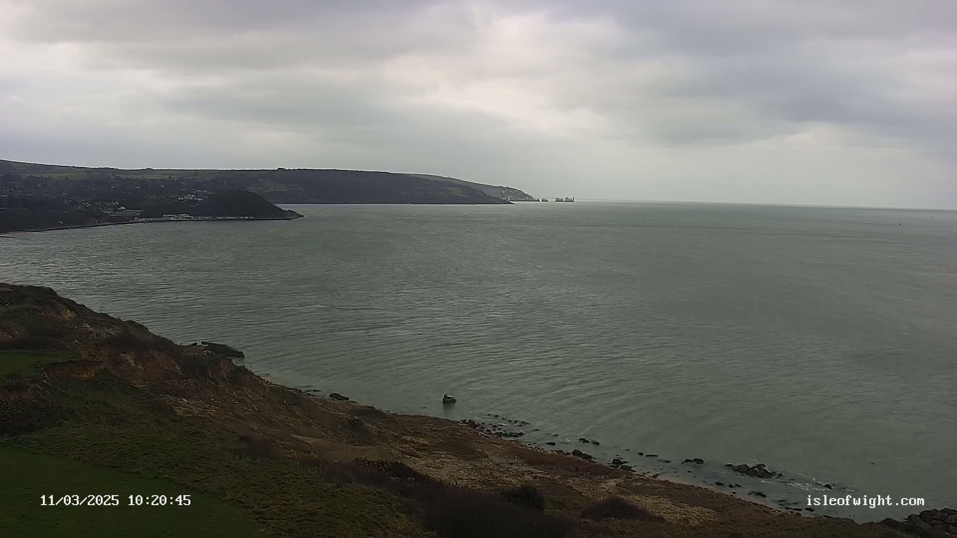A coastal view shows a calm, grey sea under an overcast sky, with a cliffside and distant land visible.