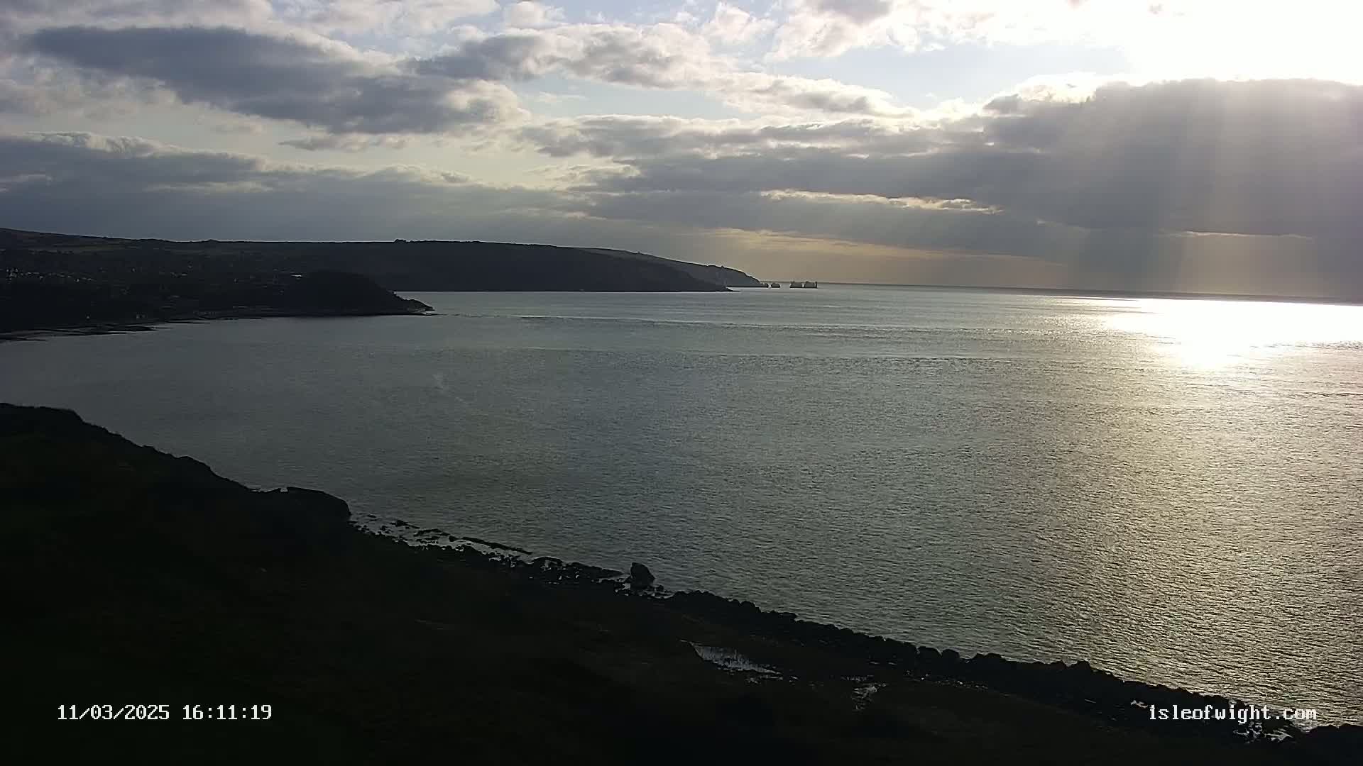 A calm body of water under a partly cloudy sky reflects sunlight, with dark landmasses visible on either side and a few distant ships.