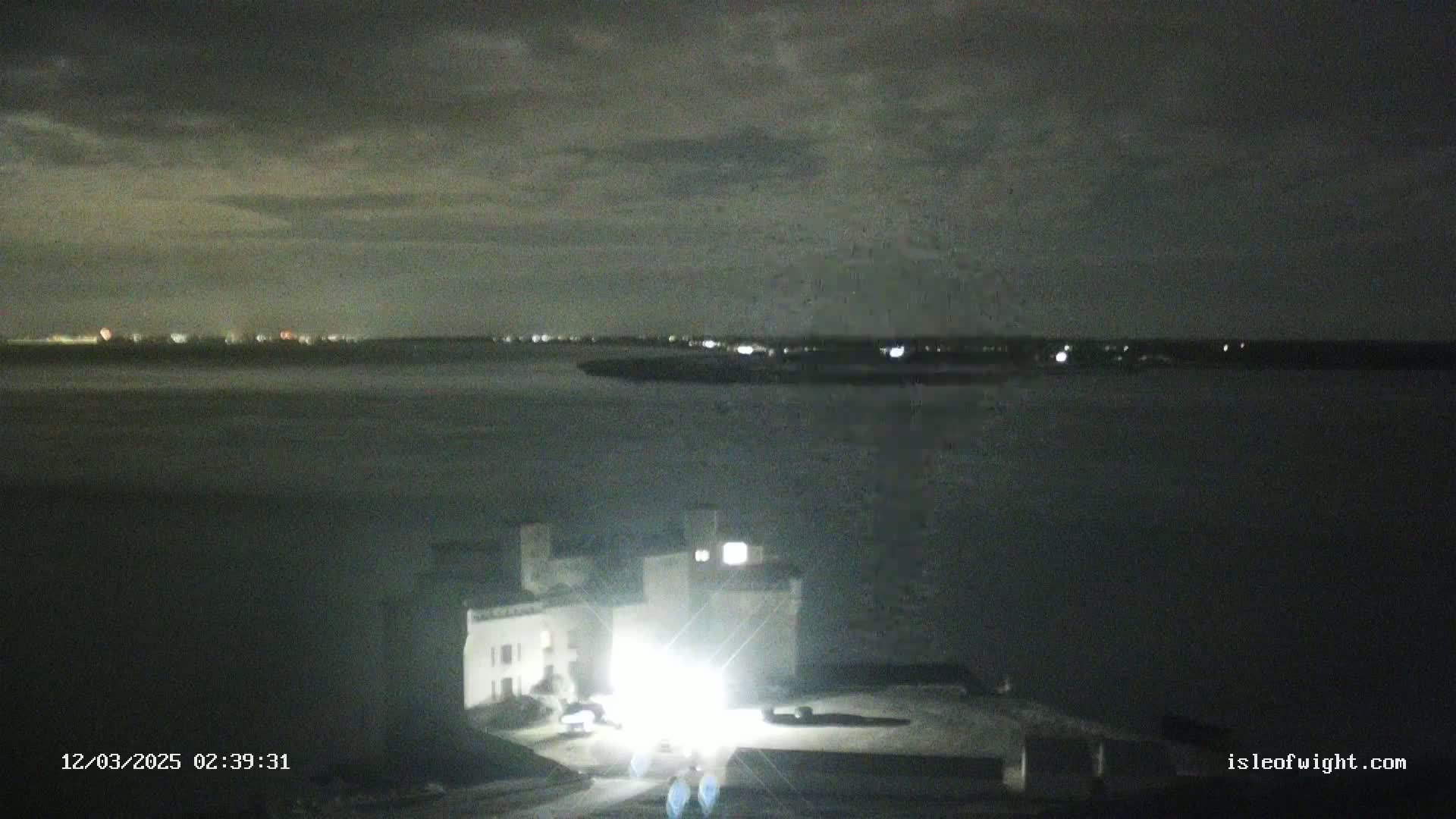 A brightly lit building sits near a dark body of water under a cloudy night sky, with distant lights visible on the opposite shore.