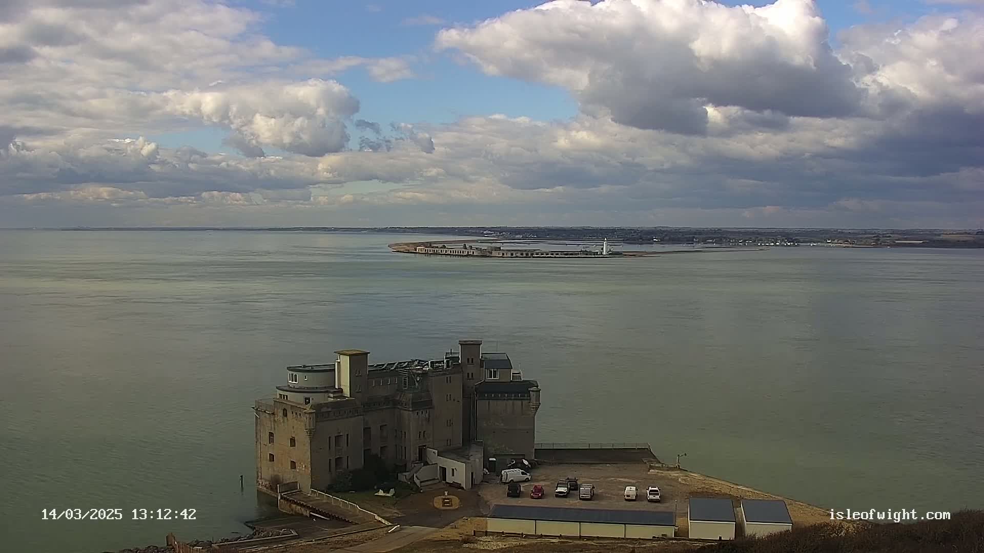 A large stone building sits on the coast overlooking a calm body of water under a partly cloudy sky.