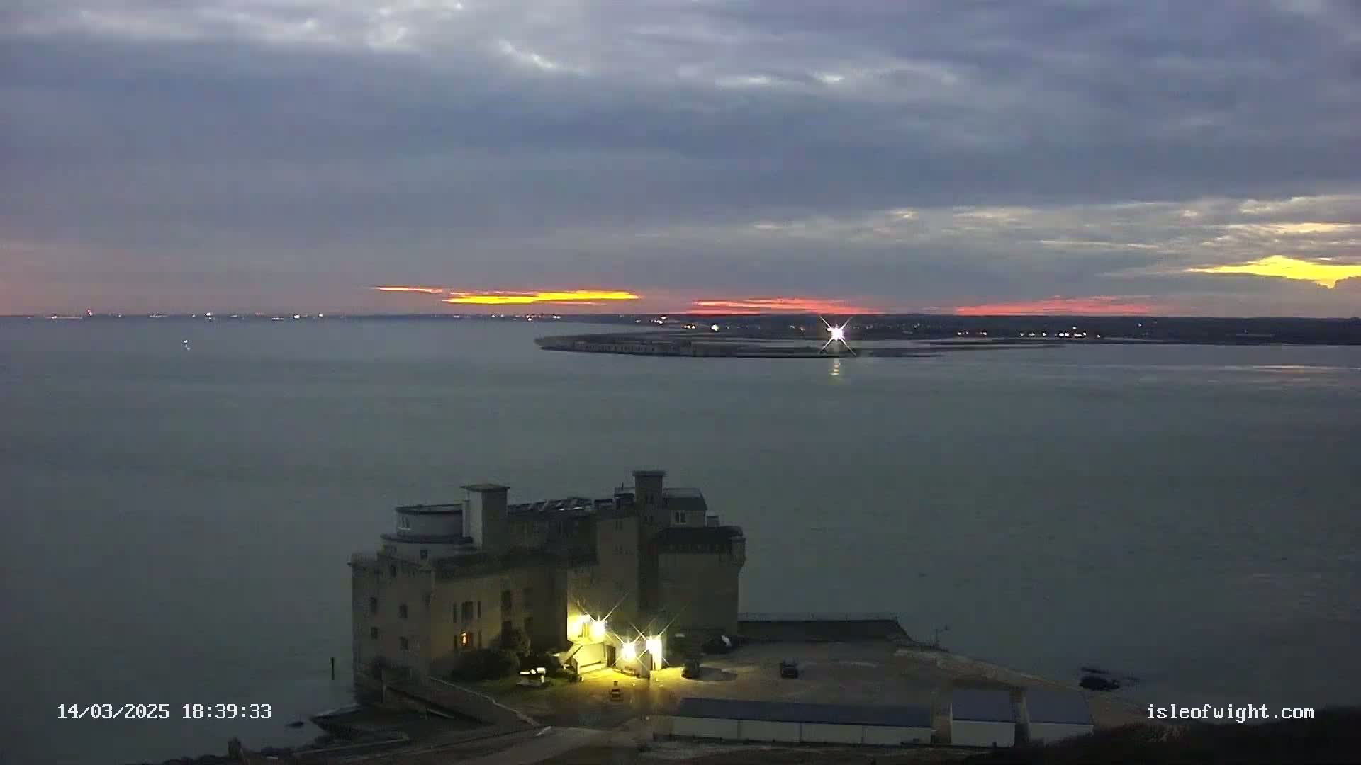A stone castle-like structure is illuminated at dusk overlooking a calm body of water with a distant sunset.