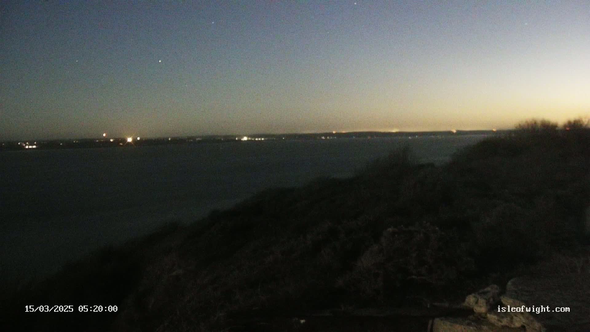 A dark, low-lit landscape overlooks a body of water with distant lights on the opposite shore under a twilight sky.