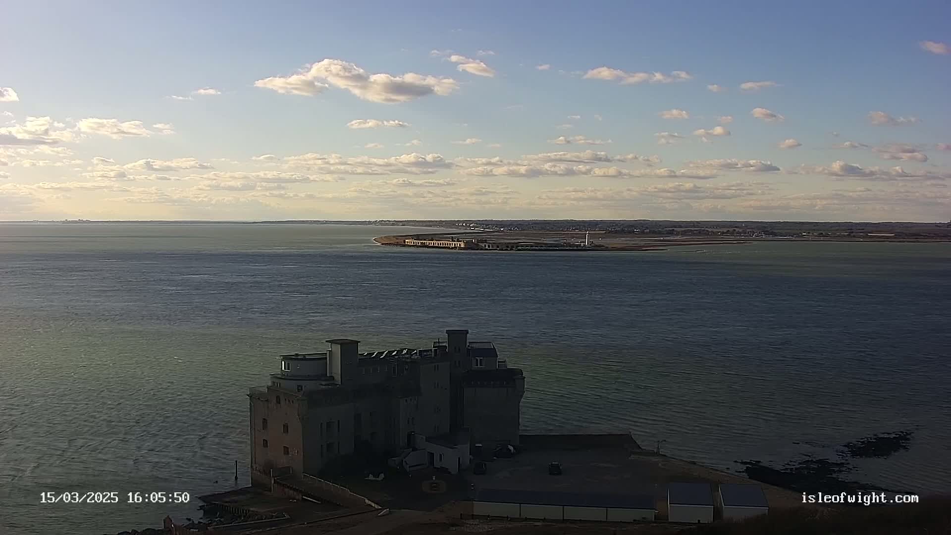 A large, stone building sits on a rocky coast overlooking a calm body of water under a partly cloudy sky.