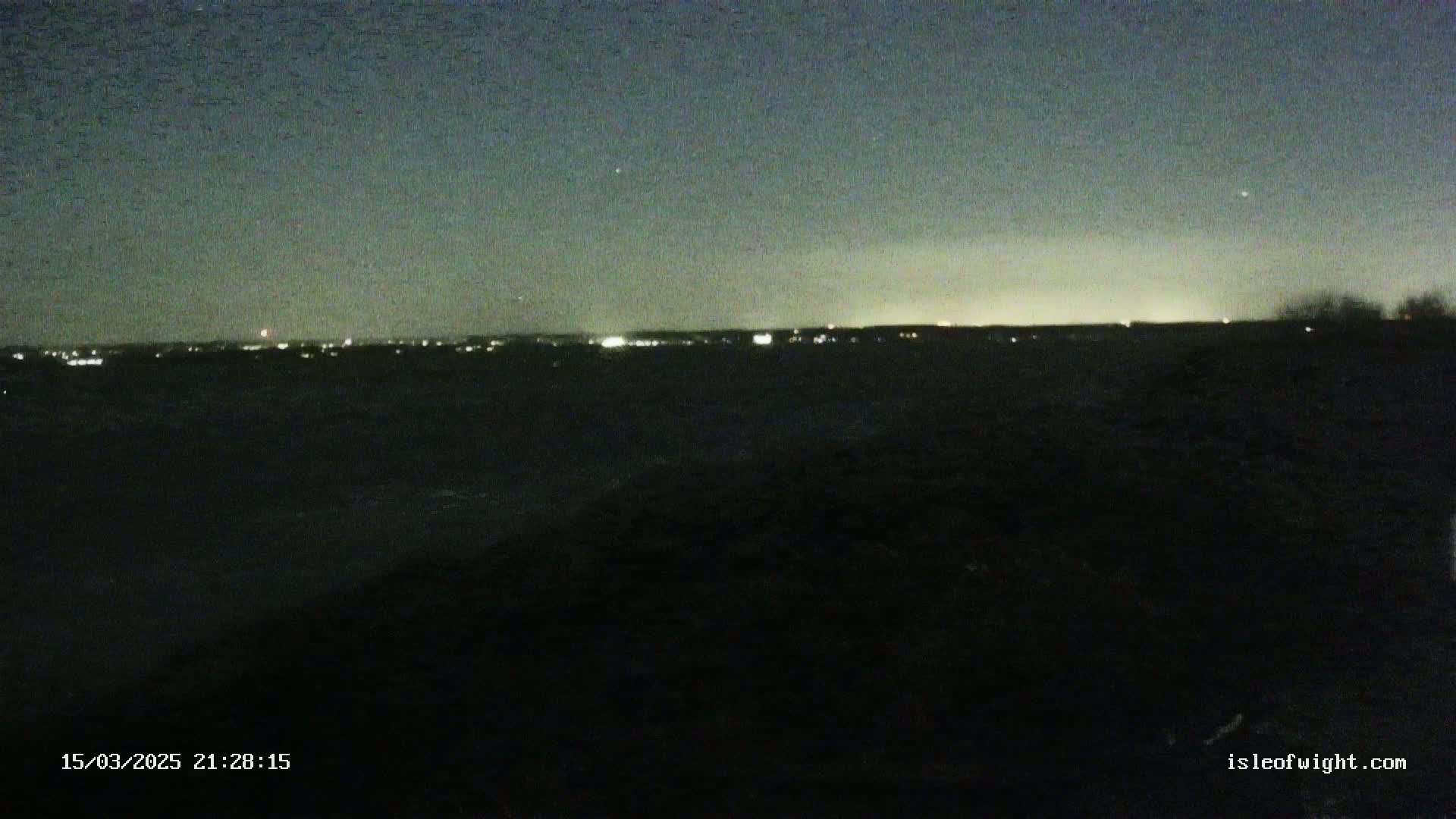 A nighttime view of a dark body of water with a distant city skyline illuminated under a clear, dark sky.