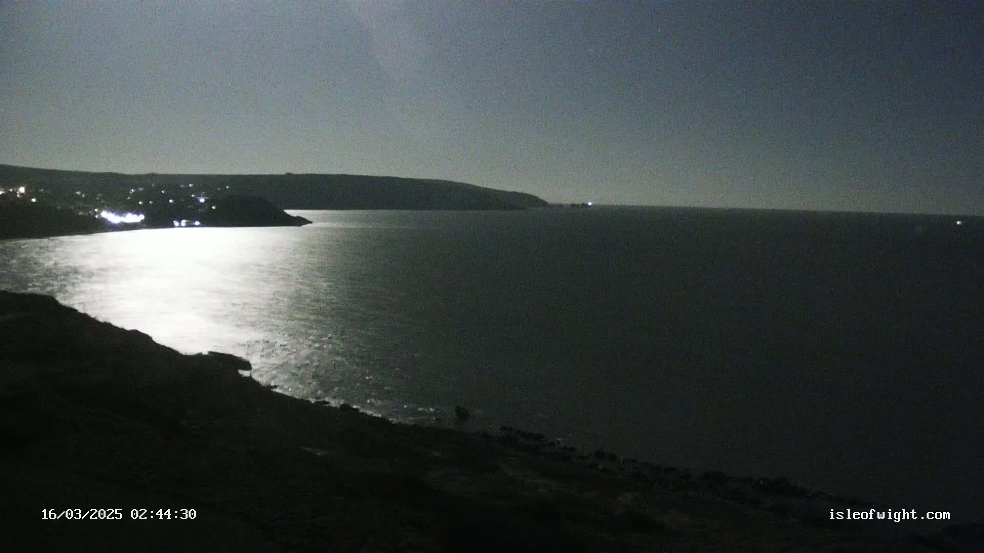 A moonlit, dark night scene shows a calm sea reflecting the light, with a distant shoreline and hills visible.