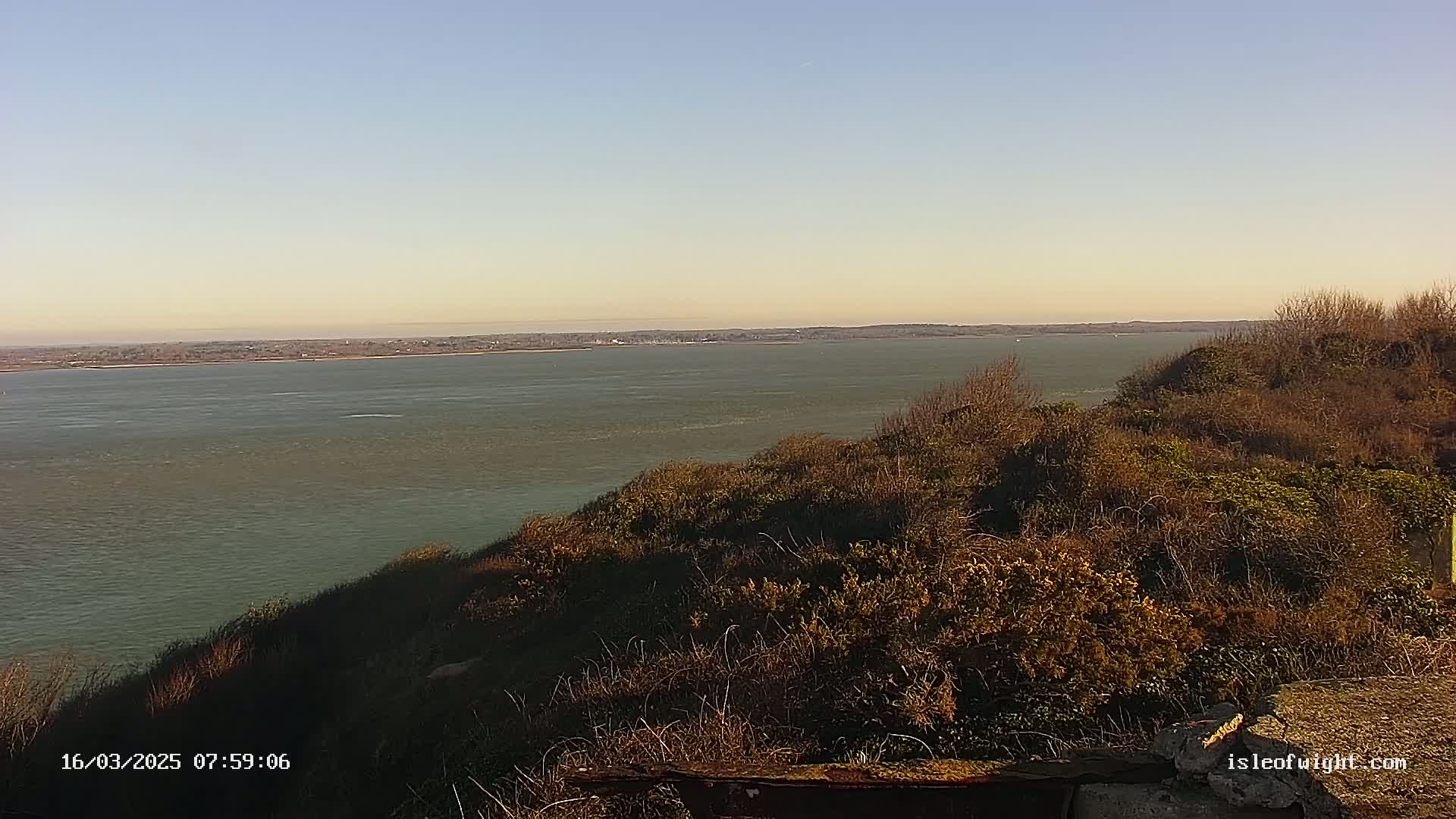 A wide shot of a calm body of water viewed from a high vantage point overlooking a scrubby, brown hillside under a clear, light-blue sky.