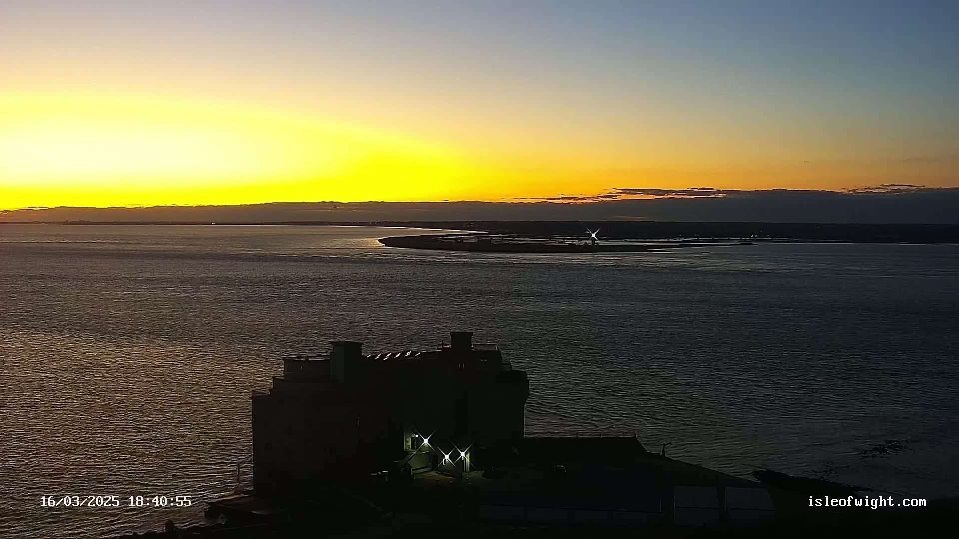 A silhouetted building with lights on sits near a body of water at sunset, with a landmass visible in the distance.