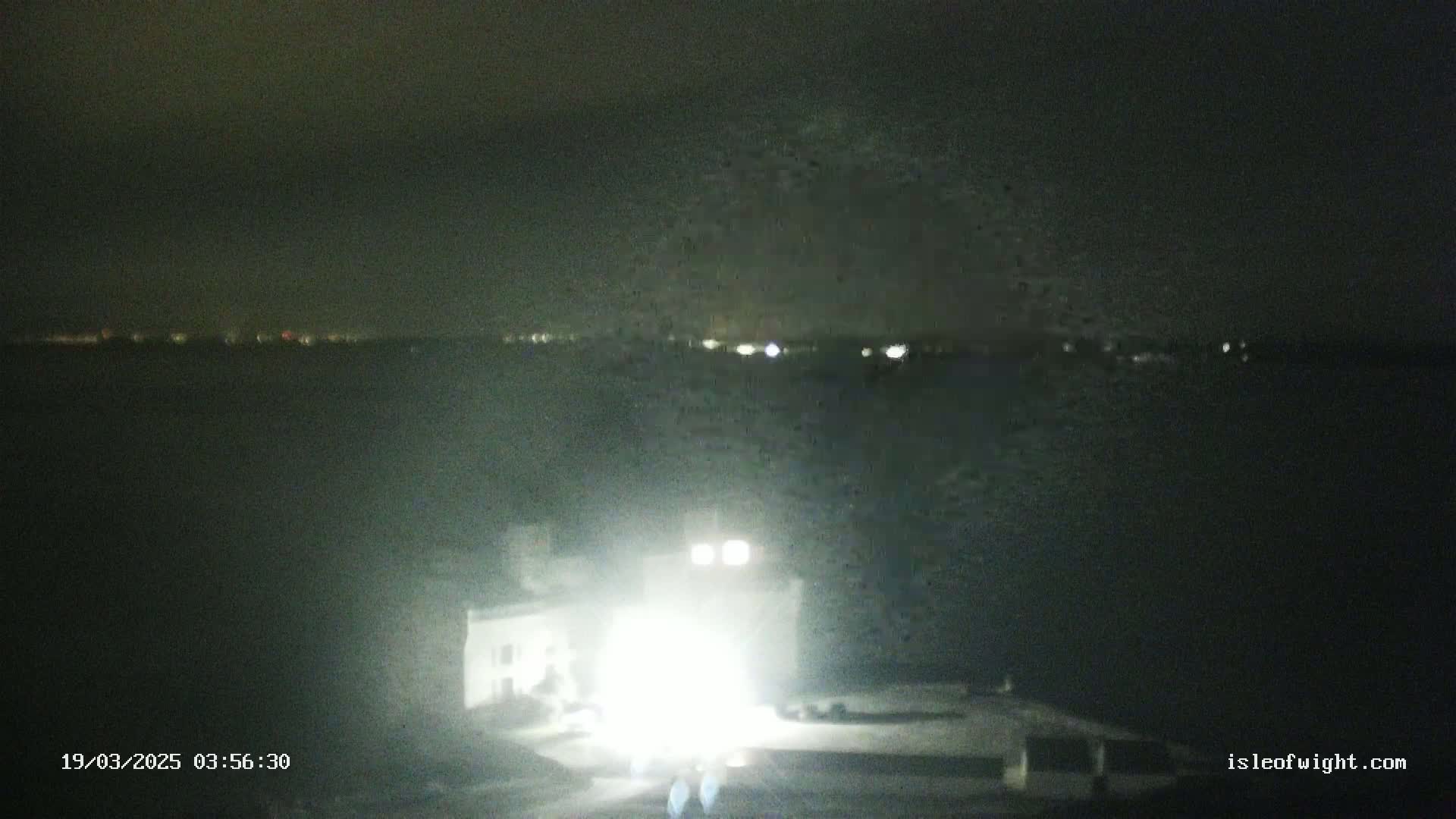 A brightly lit structure stands in the foreground against a dark, nighttime background showing a distant shoreline with faint lights.