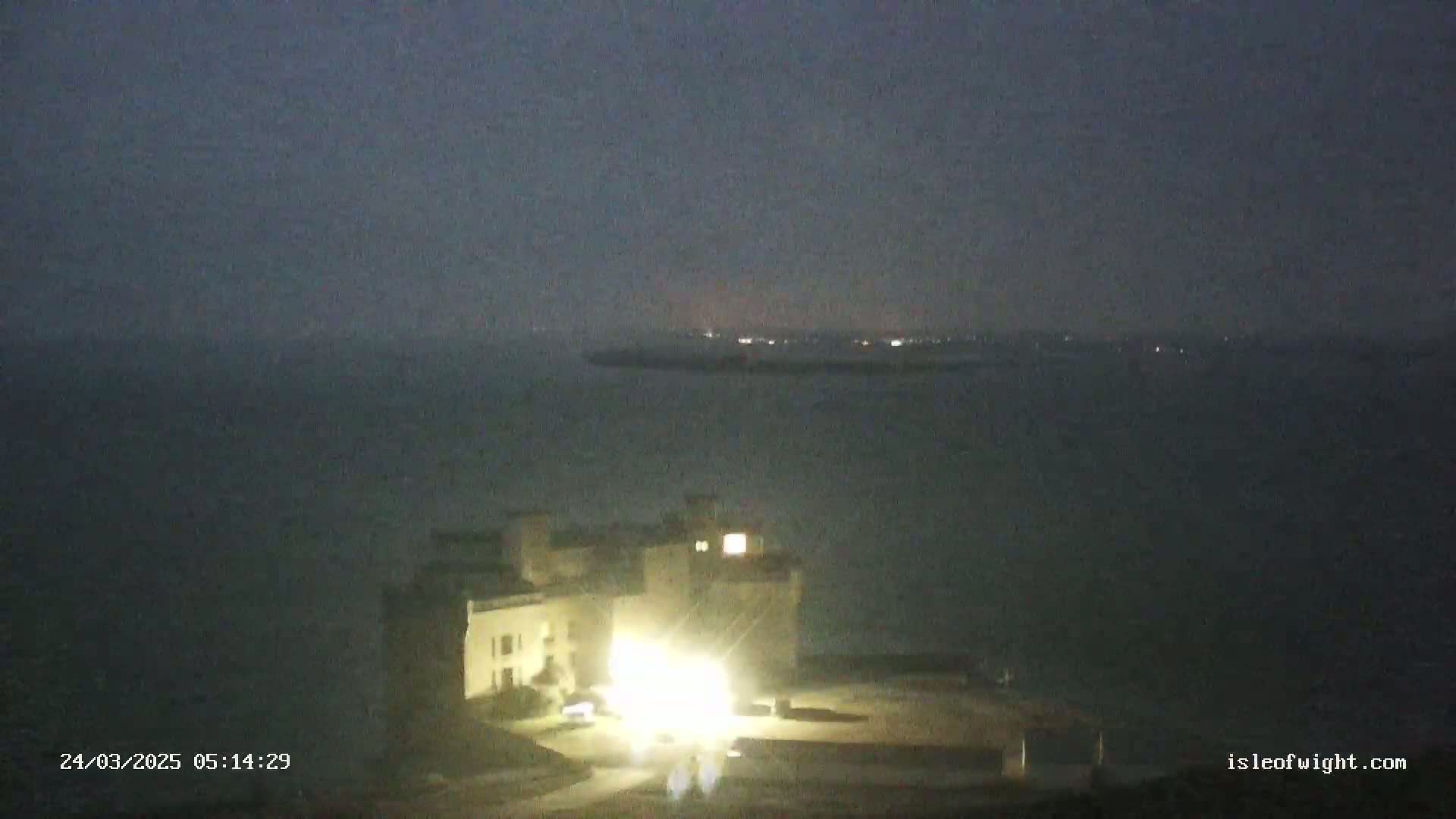 A lit building sits on the coast at night under a dark, hazy sky overlooking a calm body of water.