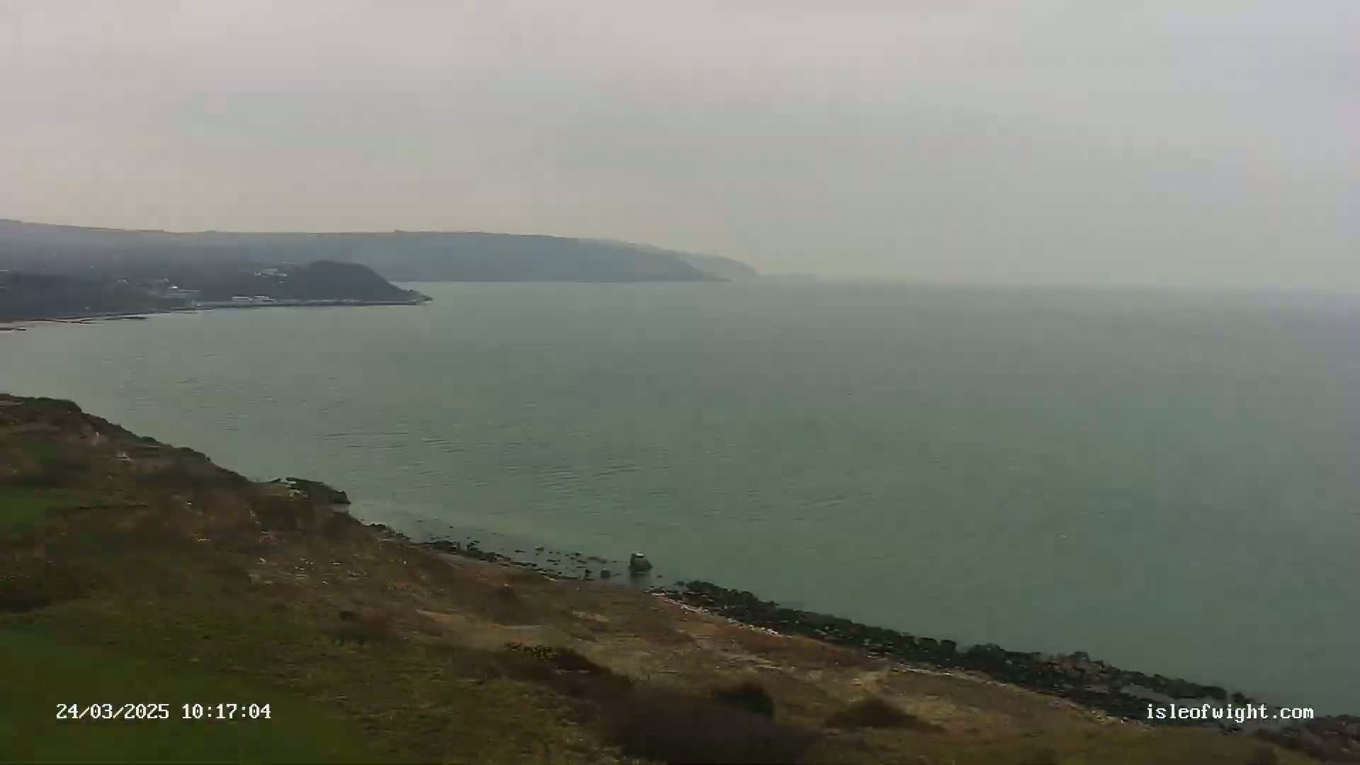 A hazy coastal scene shows a body of water with a distant coastline and a closer, grassy cliffside dropping down to a rocky shore.