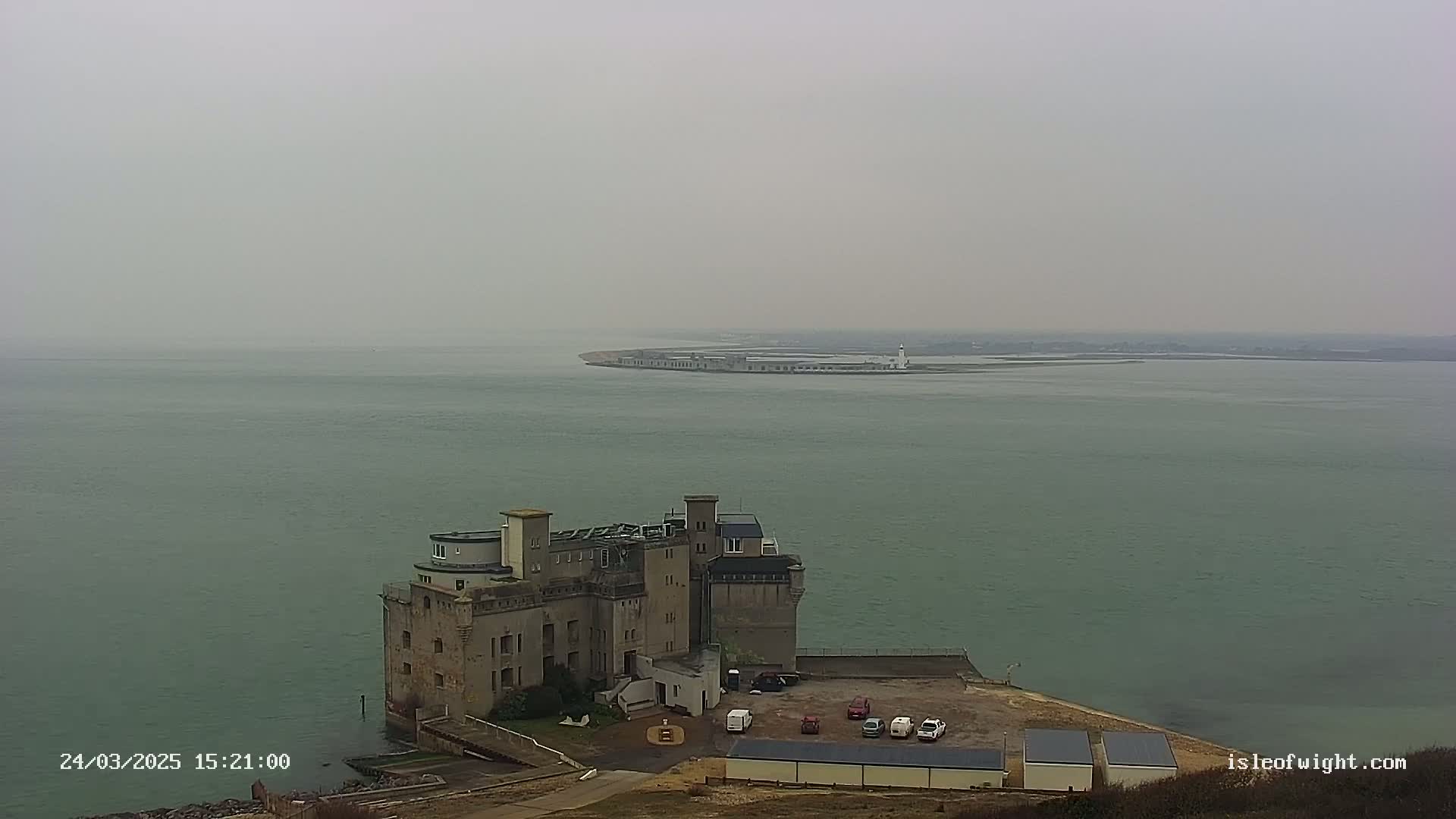 A stone building resembling a castle sits on a coast overlooking a calm, hazy body of water with a distant landmass visible.