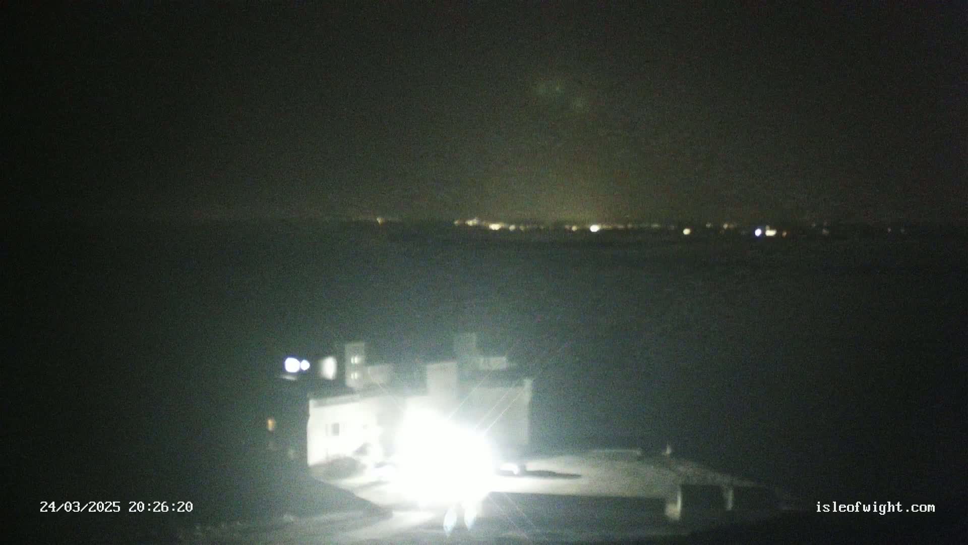 A nighttime view of a building brightly lit by headlights, with a distant line of lights on the horizon under a dark, clear sky.